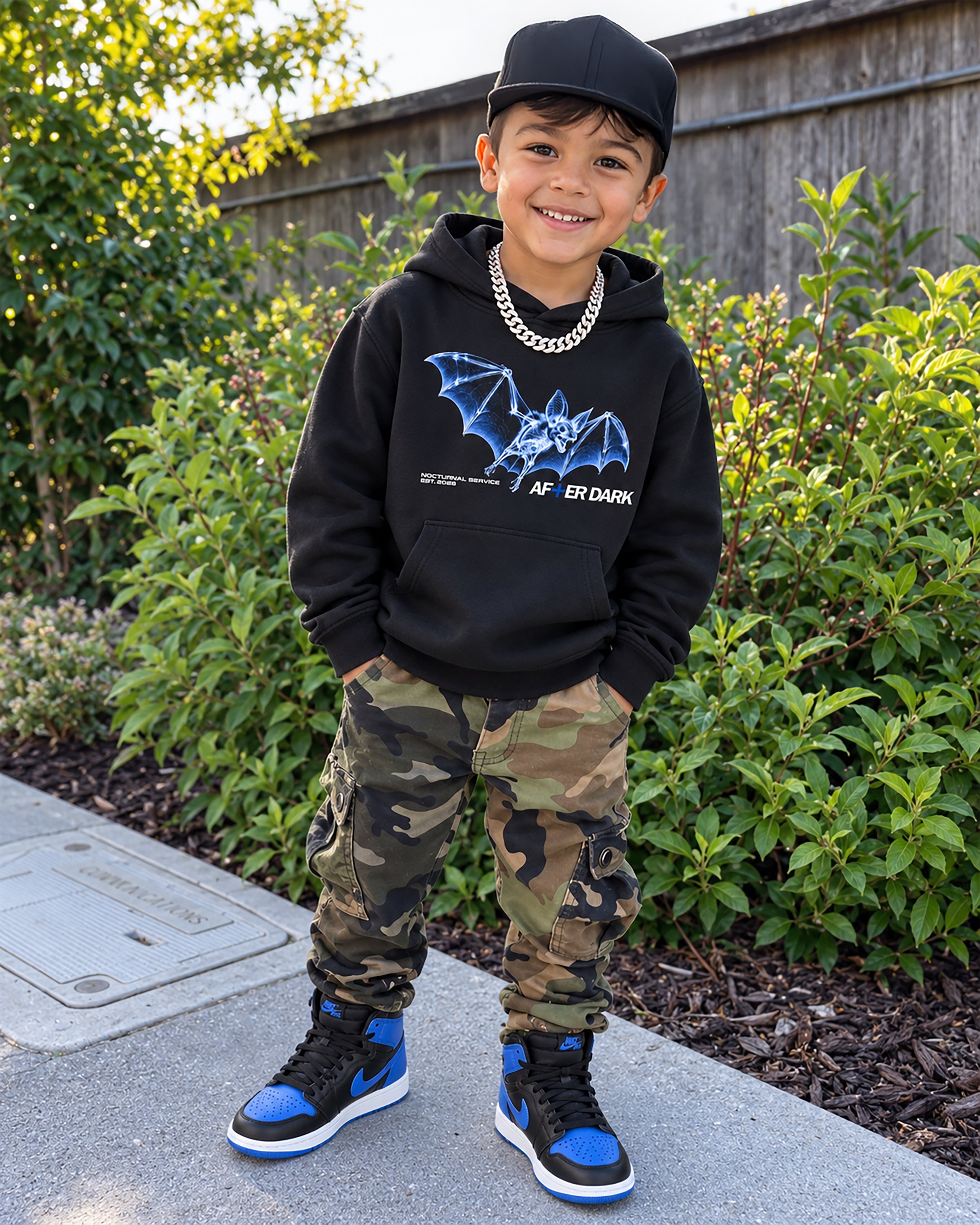 A young boy stands outdoors on a concrete path wearing a black graphic hoodie with a blue bat design, camouflage cargo pants, a black cap, and blue and black sneakers.