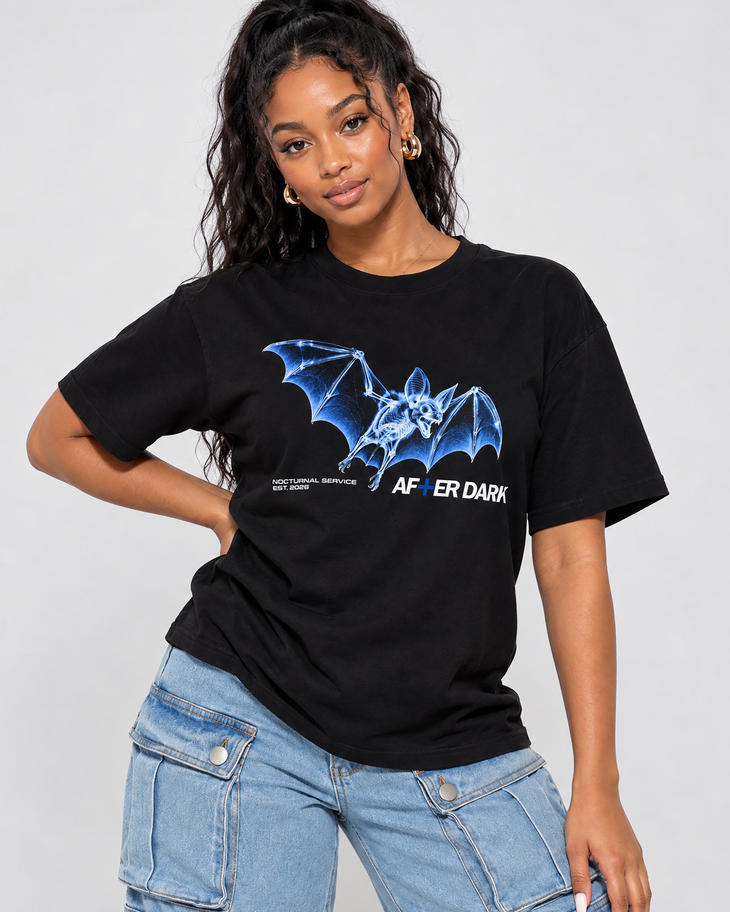 A woman wearing a black graphic t-shirt with a blue bat design and the text 'AFTER DARK' on the front, paired with light blue jeans, against a plain light gray background.