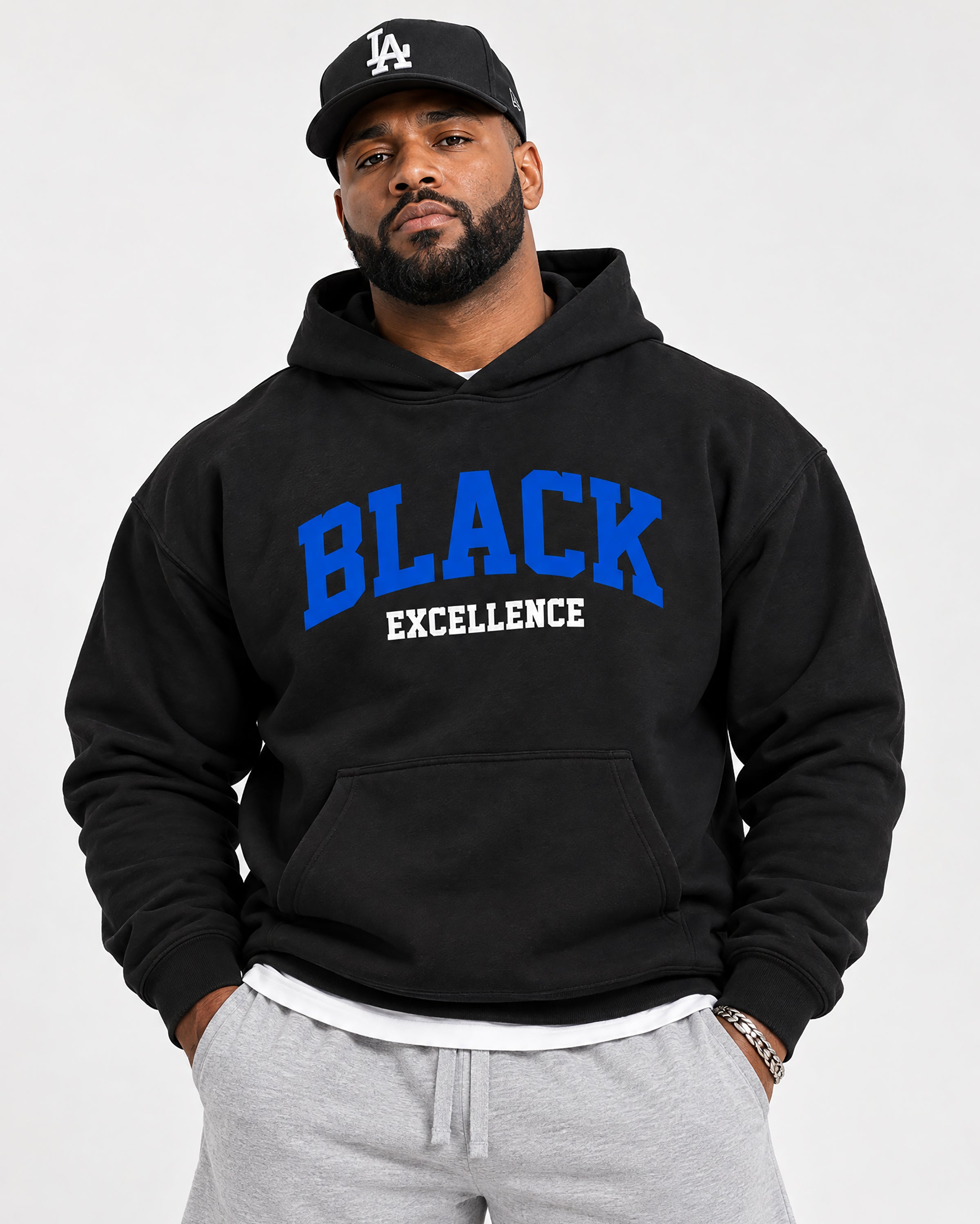 A man wearing a black pullover hoodie with 'BLACK EXCELLENCE' printed in blue and white on the front, paired with a black Los Angeles Dodgers baseball cap and light gray sweatpants, against a plain light gray background.