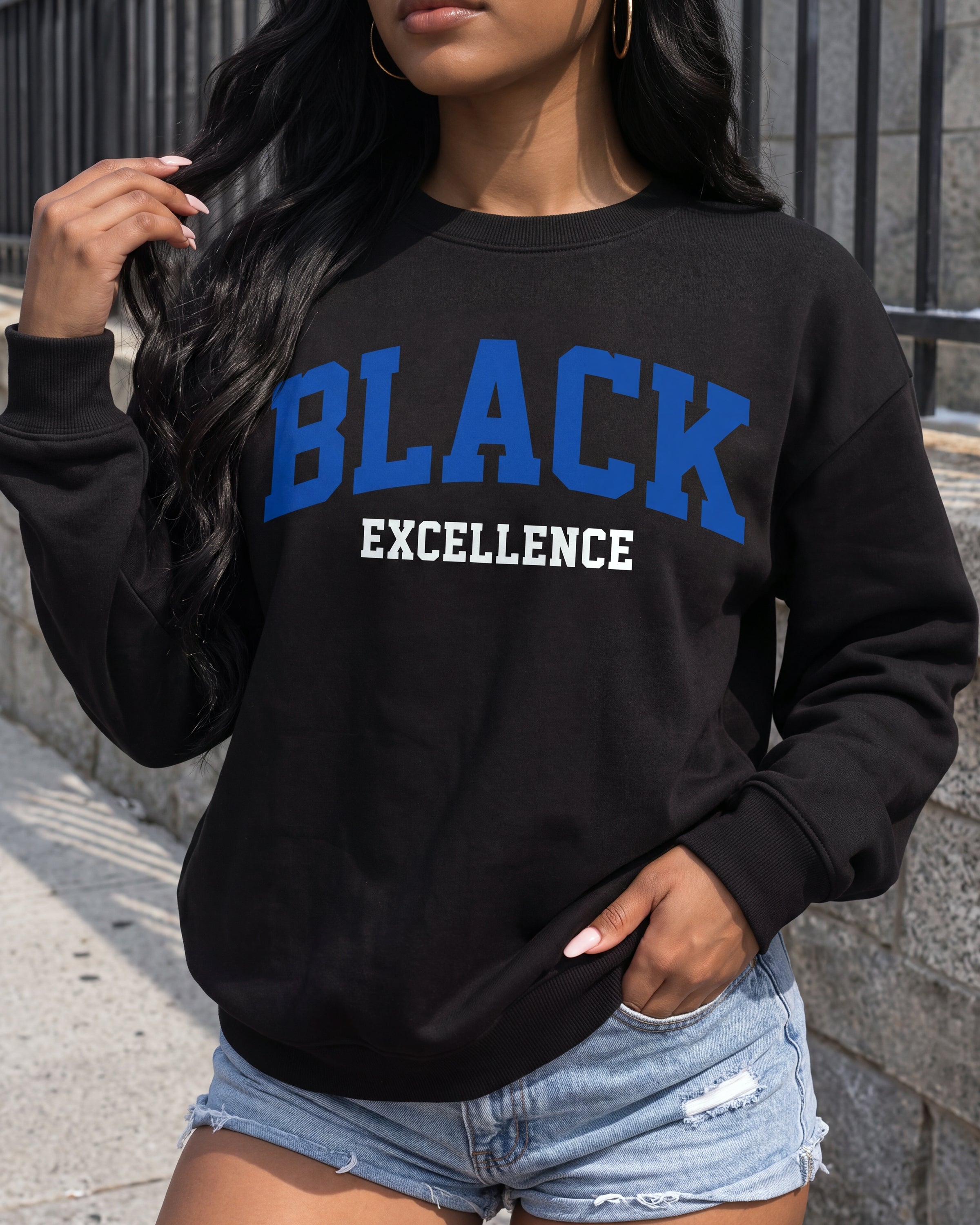 A woman wearing a black crewneck sweatshirt with 'BLACK EXCELLENCE' printed on the front in blue and white, paired with light-wash denim shorts.