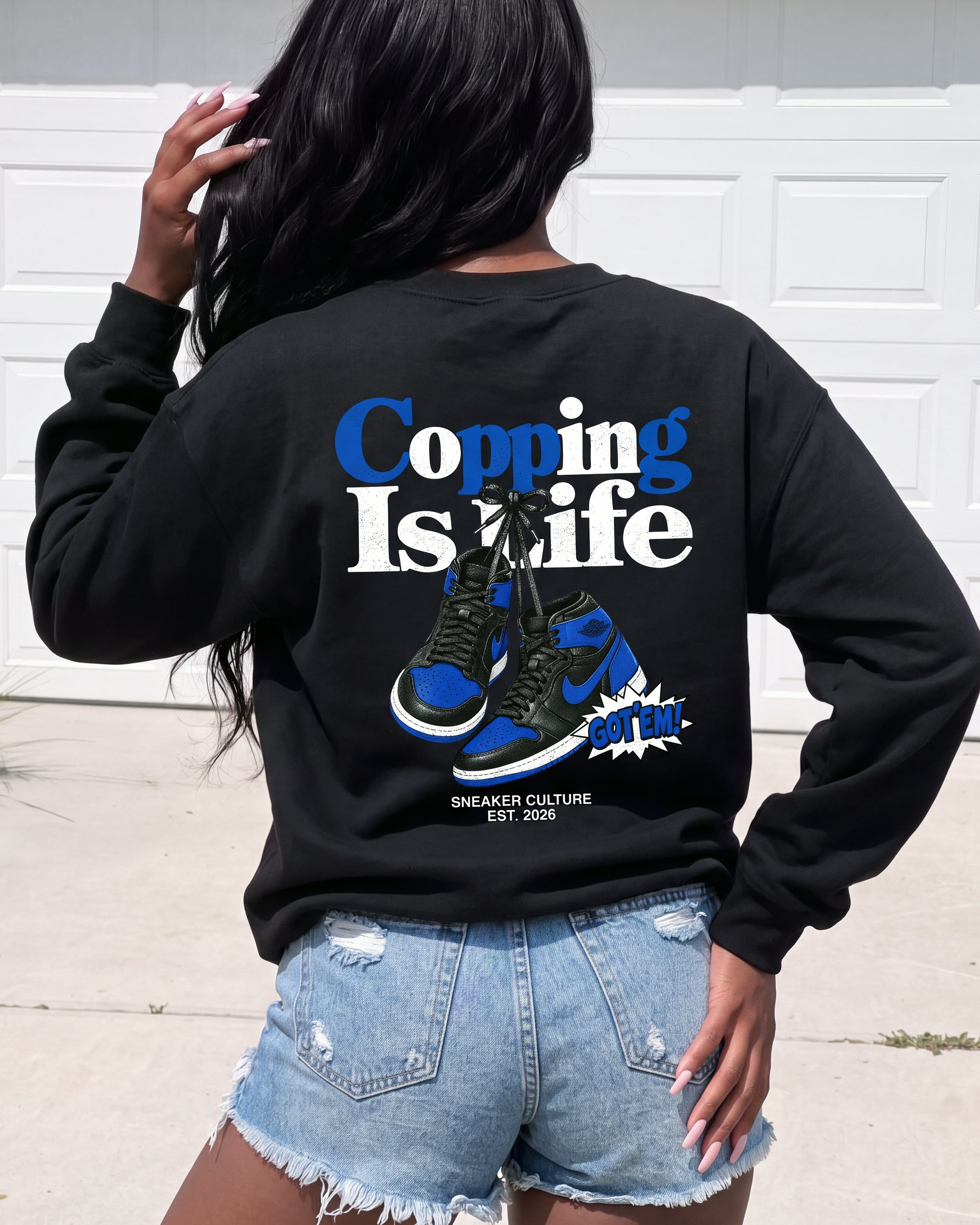 A woman shown from behind wearing a black crewneck sweatshirt with a graphic print on the back. The graphic features the text 'Copping Is Life' in blue and white, a pair of blue and black sneakers, and a small starburst that says 'BUT THE'.