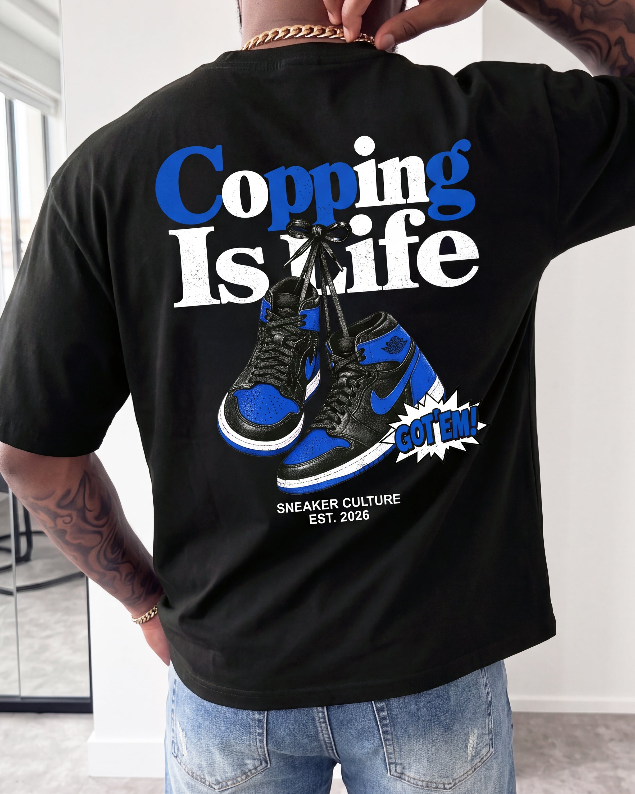 A man wearing a black t-shirt with a large graphic on the back, featuring the text 'Copping Is Life' in blue and white, a pair of blue and black sneakers, and a comic-style 'BUT TOM' starburst. The photo is taken in a lifestyle setting with a mirror visible on the left.