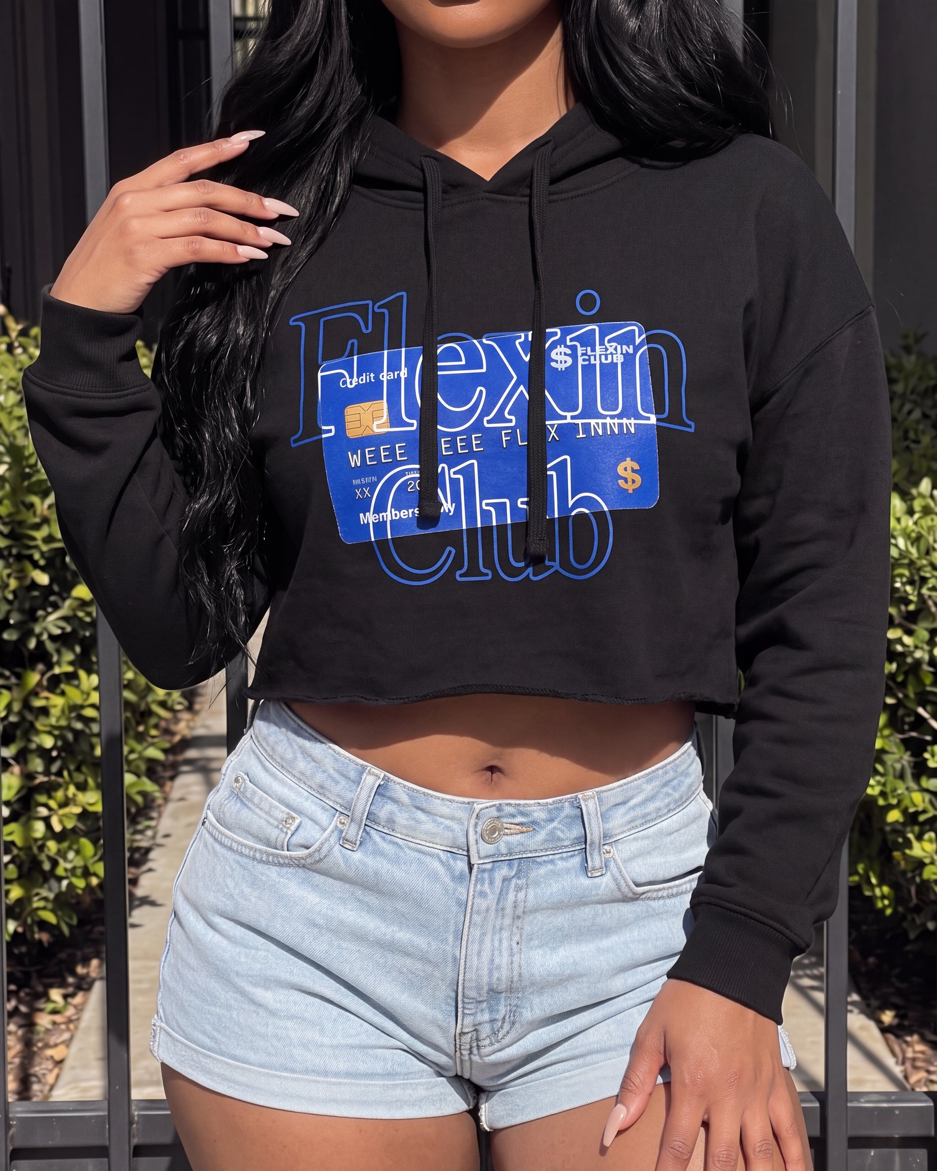 A woman wearing a black cropped hoodie with a blue credit-card-style graphic that reads 'Flexin Club' and light-wash denim shorts, standing outdoors in front of a fence and greenery.