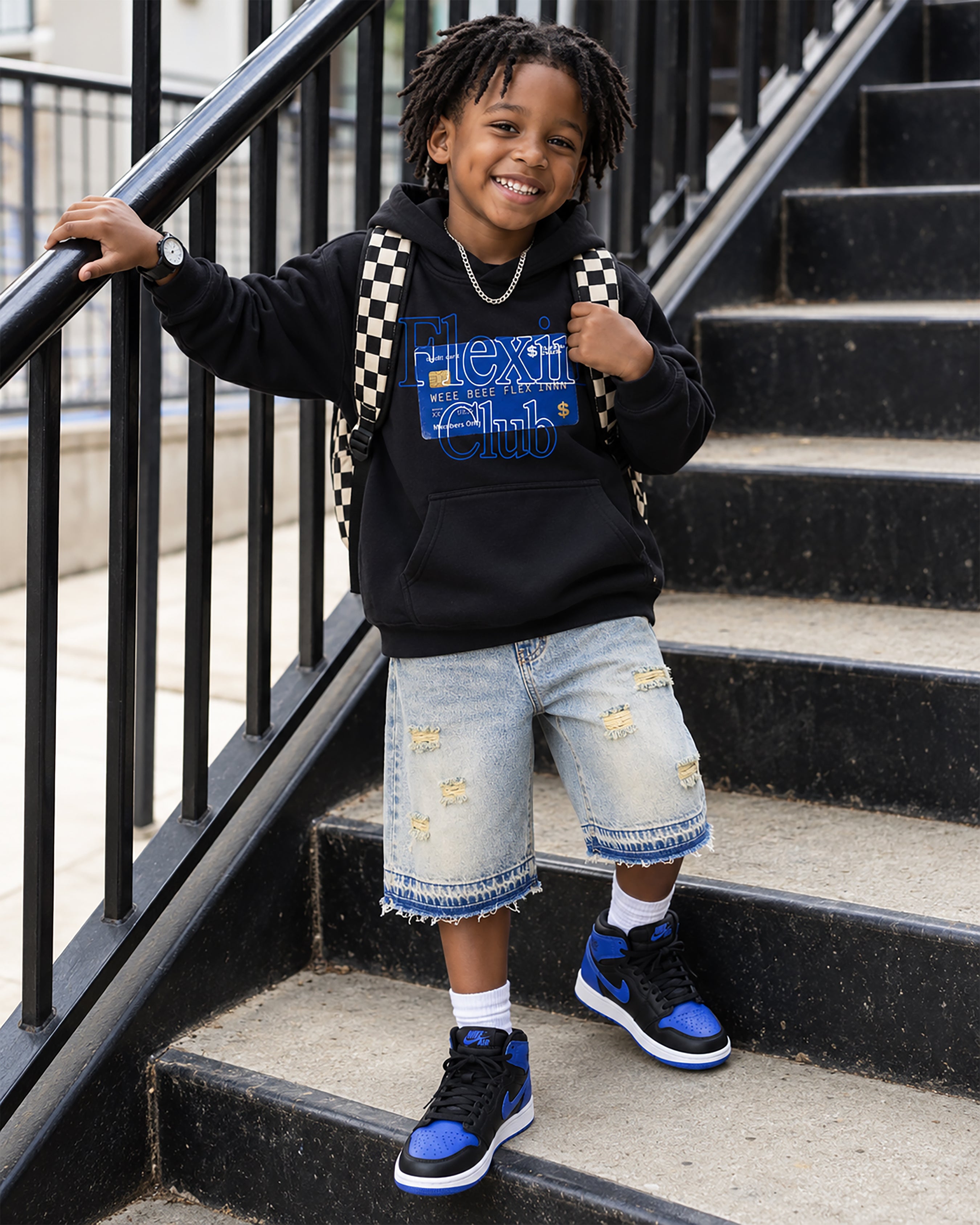 A young boy stands on outdoor concrete stairs holding a black metal railing, wearing a black hoodie with a blue and white graphic that reads 'Next Gen Club', light-wash distressed denim shorts, white socks, and blue and black sneakers. He is also wearing a black-and-white checkered backpack.