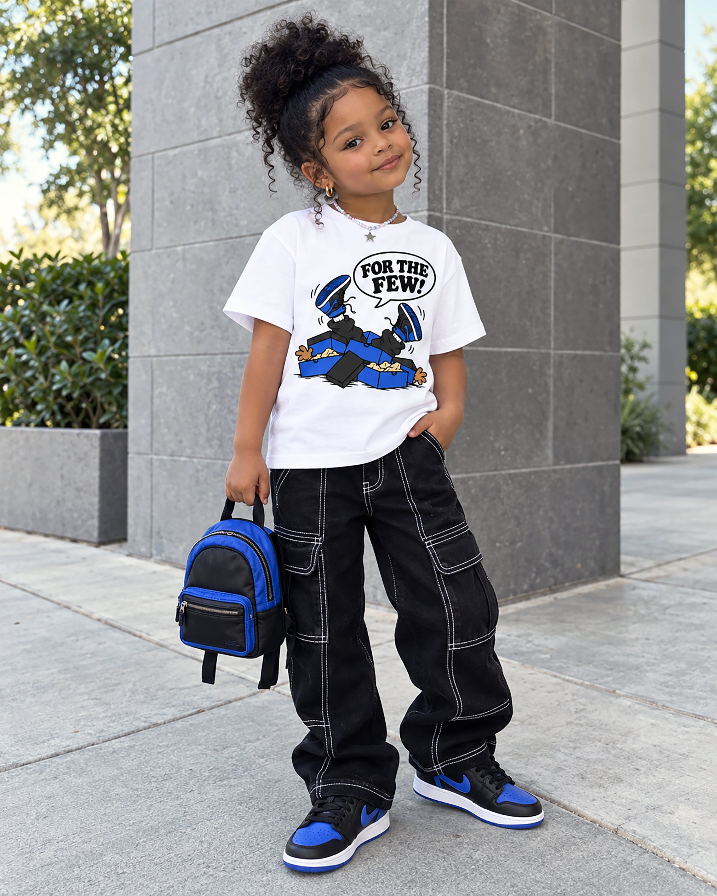 A young girl stands outdoors in front of a gray stone column, wearing a white graphic T-shirt with a cartoon character and the text 'FOR THE FEW!', black cargo pants with white contrast stitching, blue and black sneakers, and carrying a small blue and black backpack.