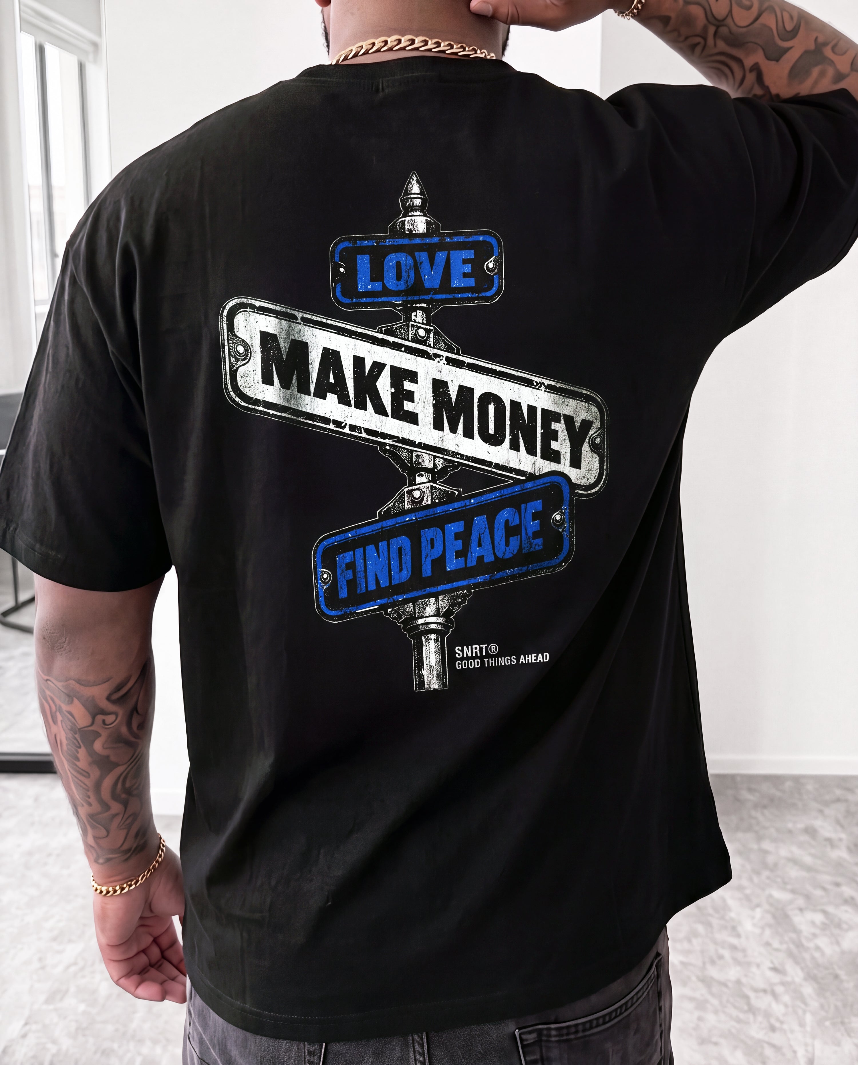 A man wearing a black t-shirt, viewed from the back. The shirt features a graphic print of three street signs pointing in opposite directions with the words 'LOVE', 'MAKE MONEY', and 'FIND PEACE' in blue and white, with 'SHUT UP' printed below.