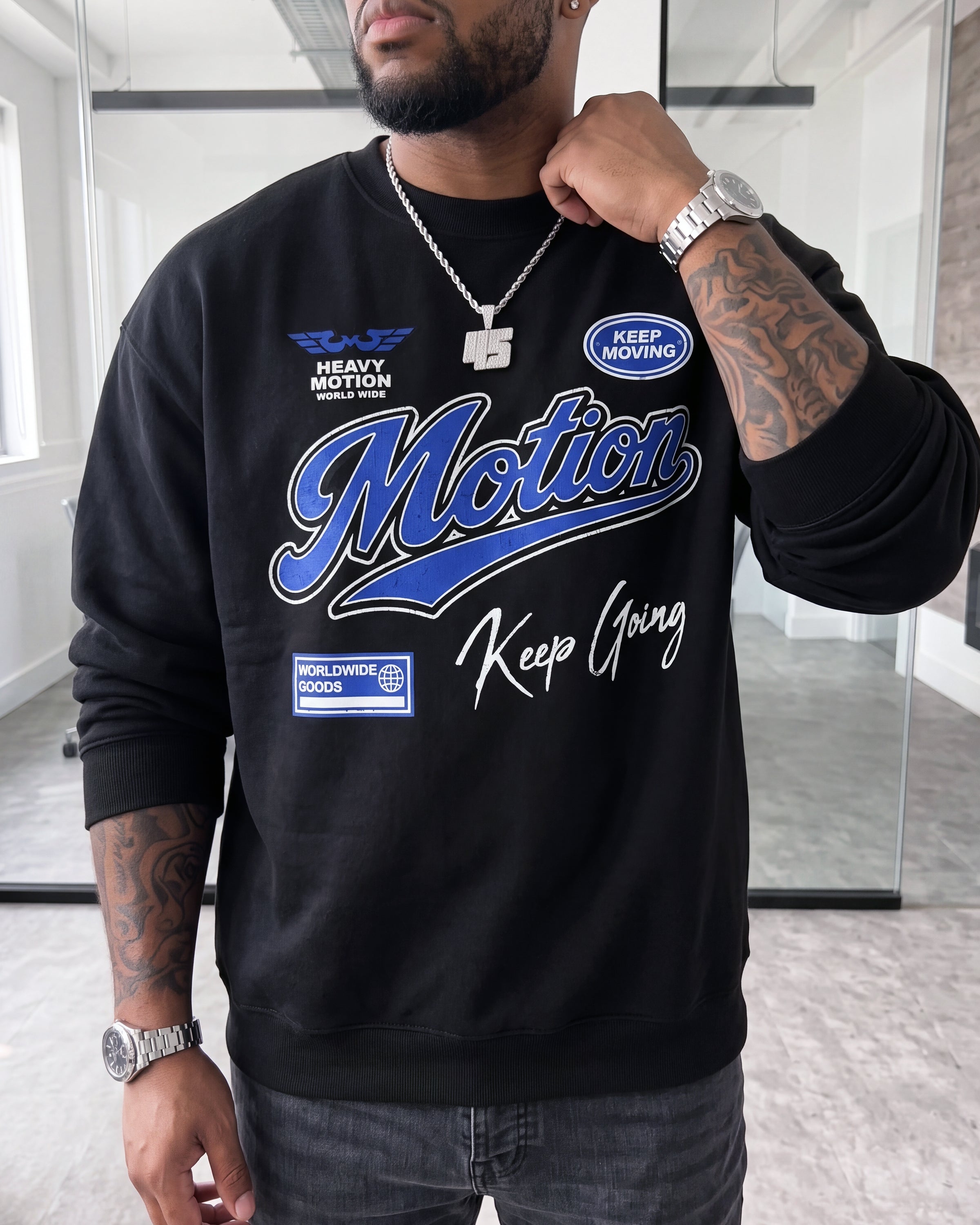 A man wearing a black crewneck sweatshirt with blue and white graphic text reading 'Motion' and 'Keep Going' on the front, paired with gray jeans and silver jewelry, in an indoor studio setting.