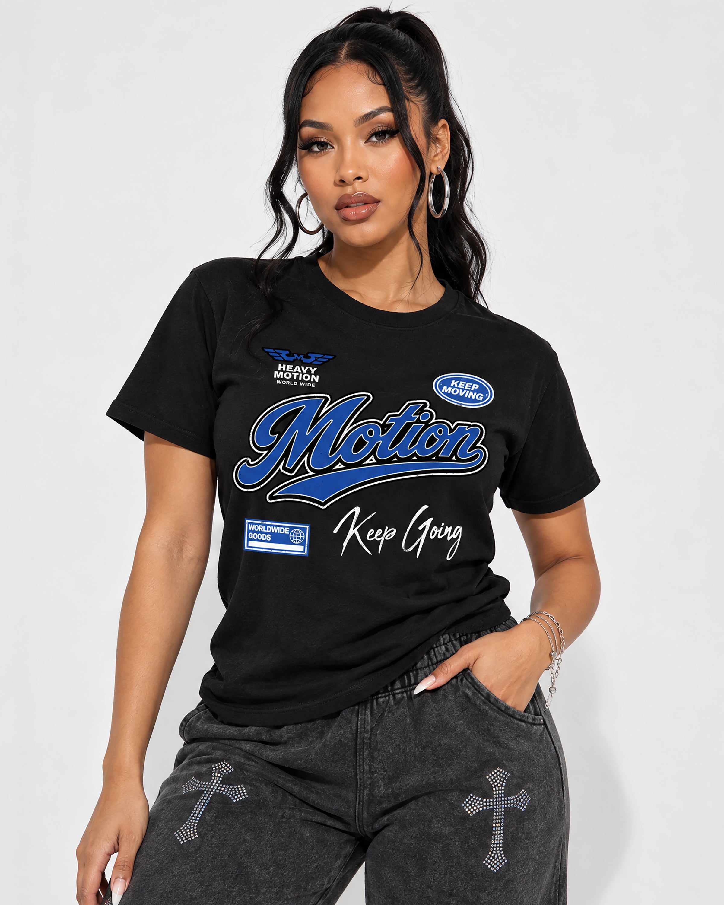 A woman modeling a black graphic t-shirt with blue and white 'Motion Keep Goong' text, paired with gray jeans featuring rhinestone cross details, against a light gray studio background.