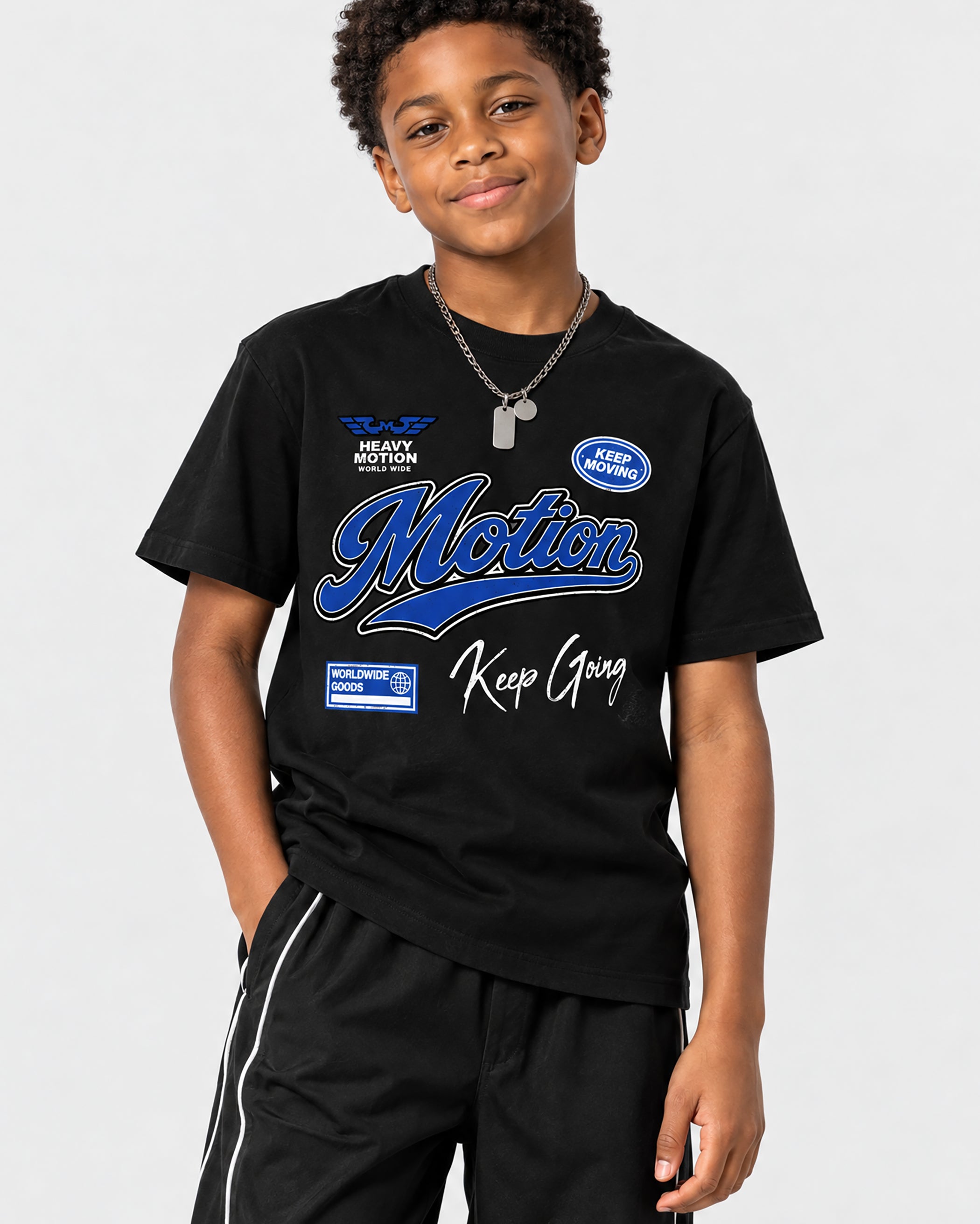 A young boy wearing a black graphic t-shirt with blue and white 'Motion Keep Goong' text, paired with black track pants, against a light gray studio background.