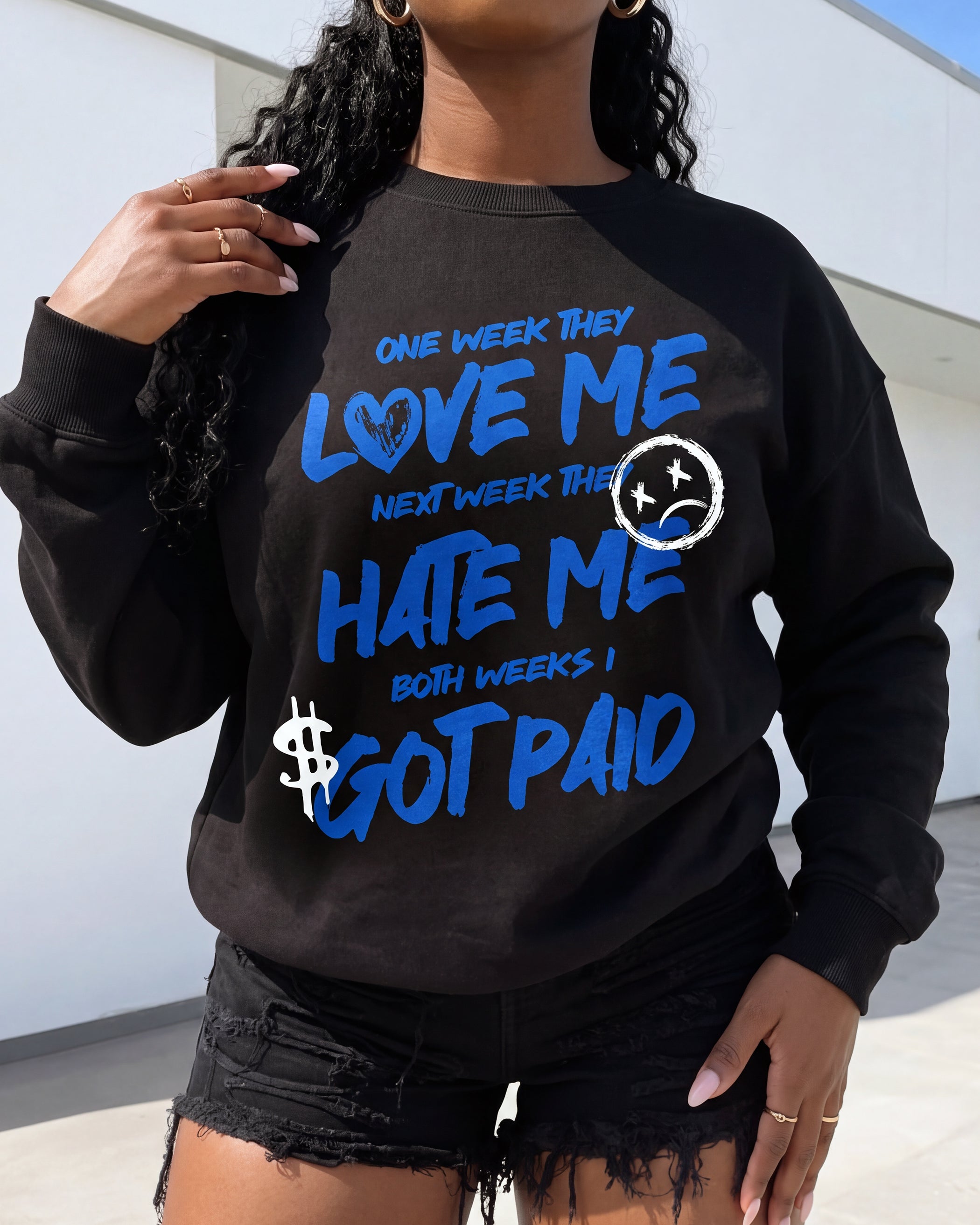 A woman wearing a black crewneck sweatshirt with a blue and white graphic text that reads 'ONE WEEK THEY LOVE ME NEXT WEEK THEY HATE ME BOTH WEEKS I GOT PAID' along with a dollar sign and sad face graphic, paired with black distressed denim shorts.