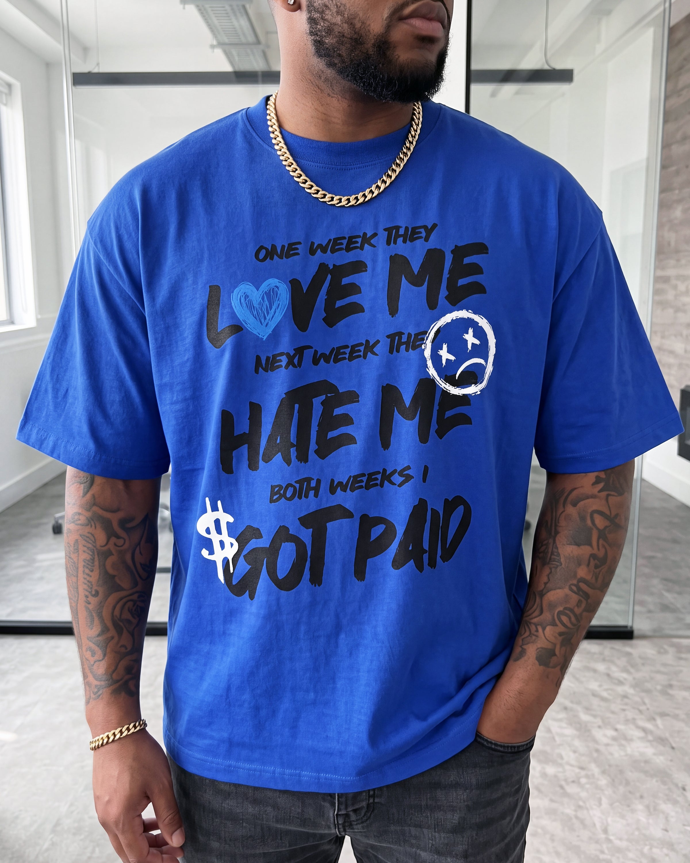 A man wearing a bright blue graphic t-shirt with the text 'ONE WEEK THEY LOVE ME NEXT WEEK THEY HATE ME BOTH WEEKS I GOT PAID' in black and blue lettering, paired with dark gray jeans and a gold chain necklace.