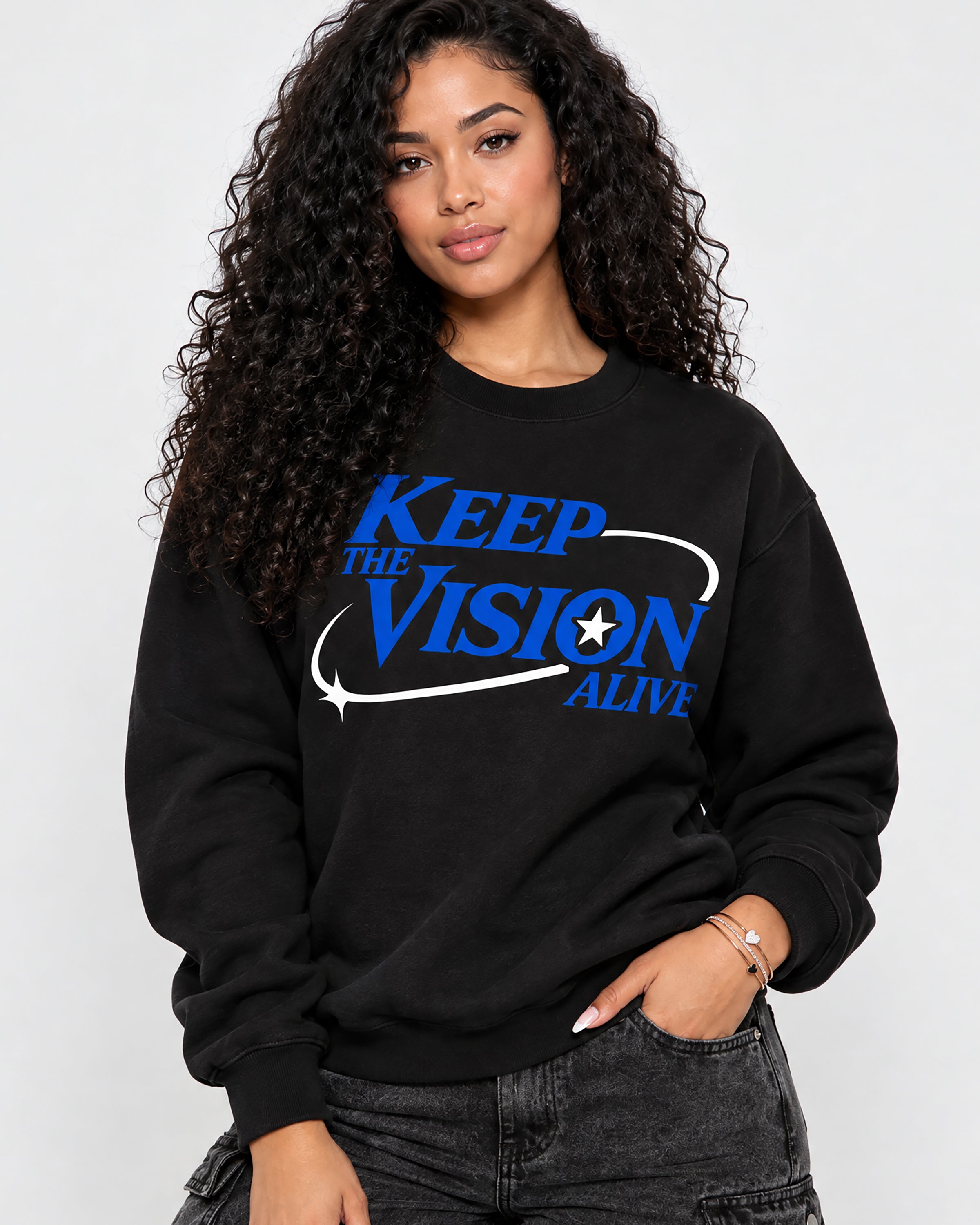 A woman with long, curly dark hair wearing a black crewneck sweatshirt with a blue and white graphic that reads 'KEEP THE VISION ALIVE' and dark gray jeans, posing against a light gray studio background.