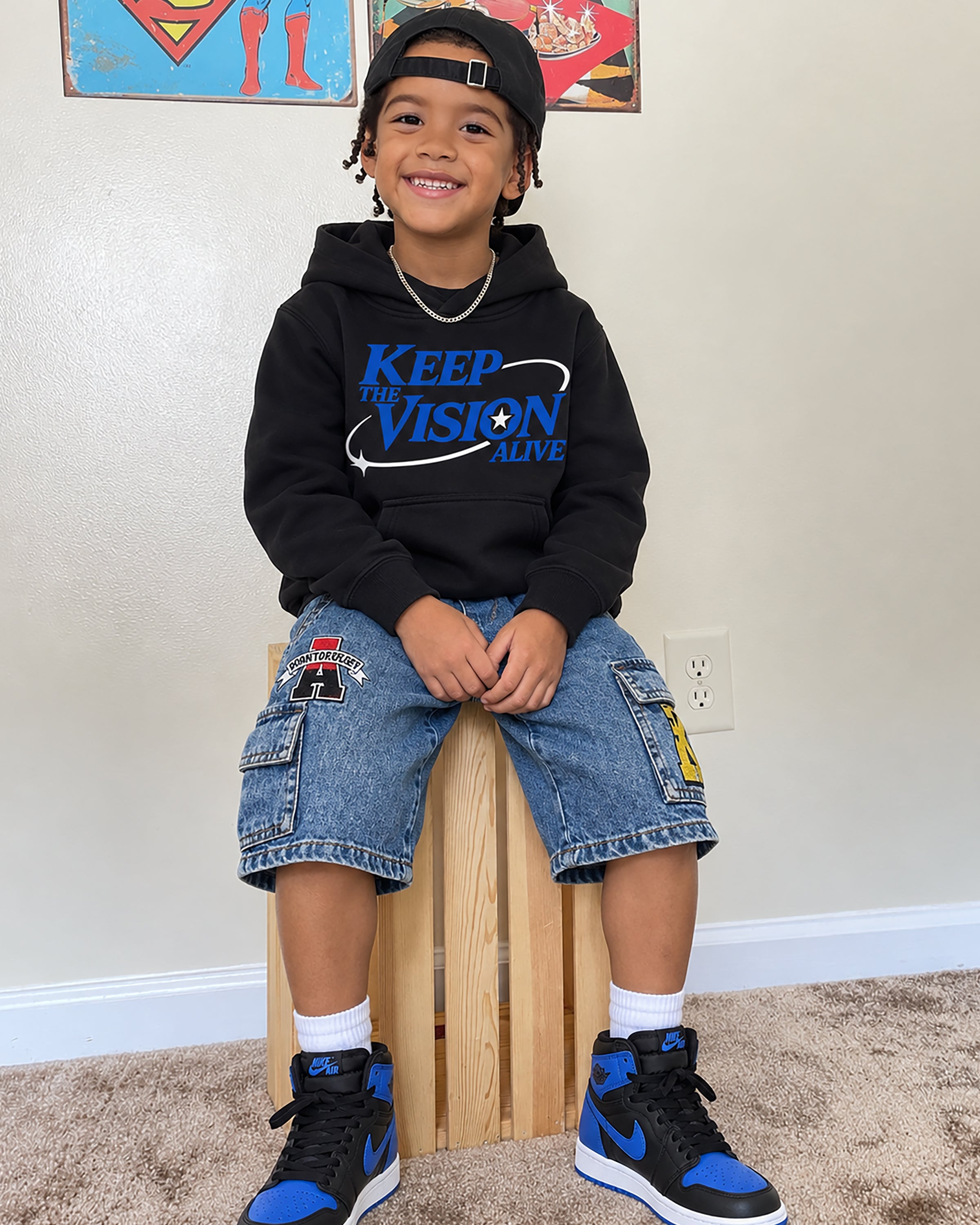 A young boy wearing a black hoodie with blue graphic text, denim cargo shorts, and blue and black sneakers, sitting on a wooden block in a home setting.