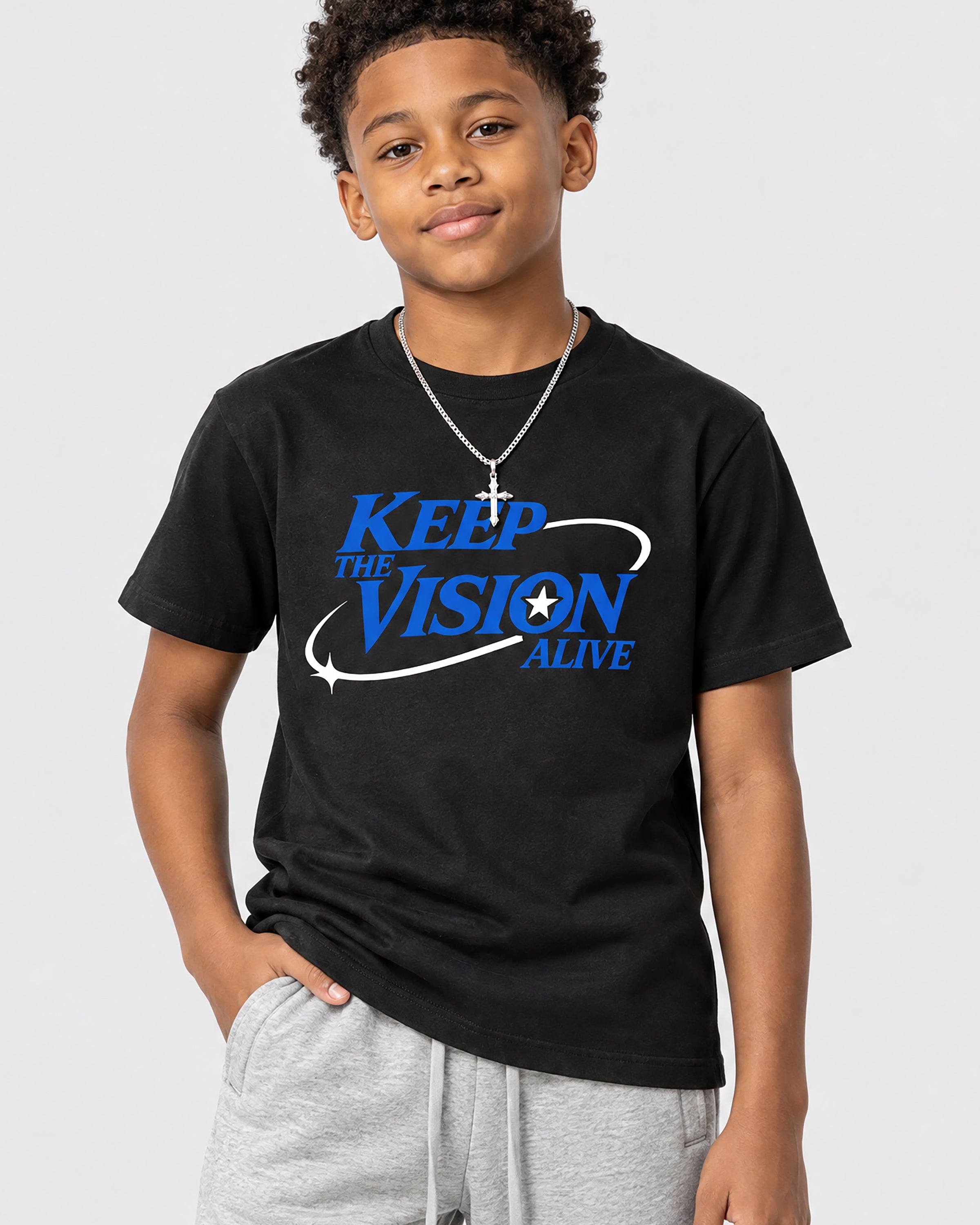 A young boy wearing a black graphic t-shirt with the text 'KEEP THE VISION ALIVE' in blue and white, paired with gray sweatpants and a silver cross necklace, against a light gray background.