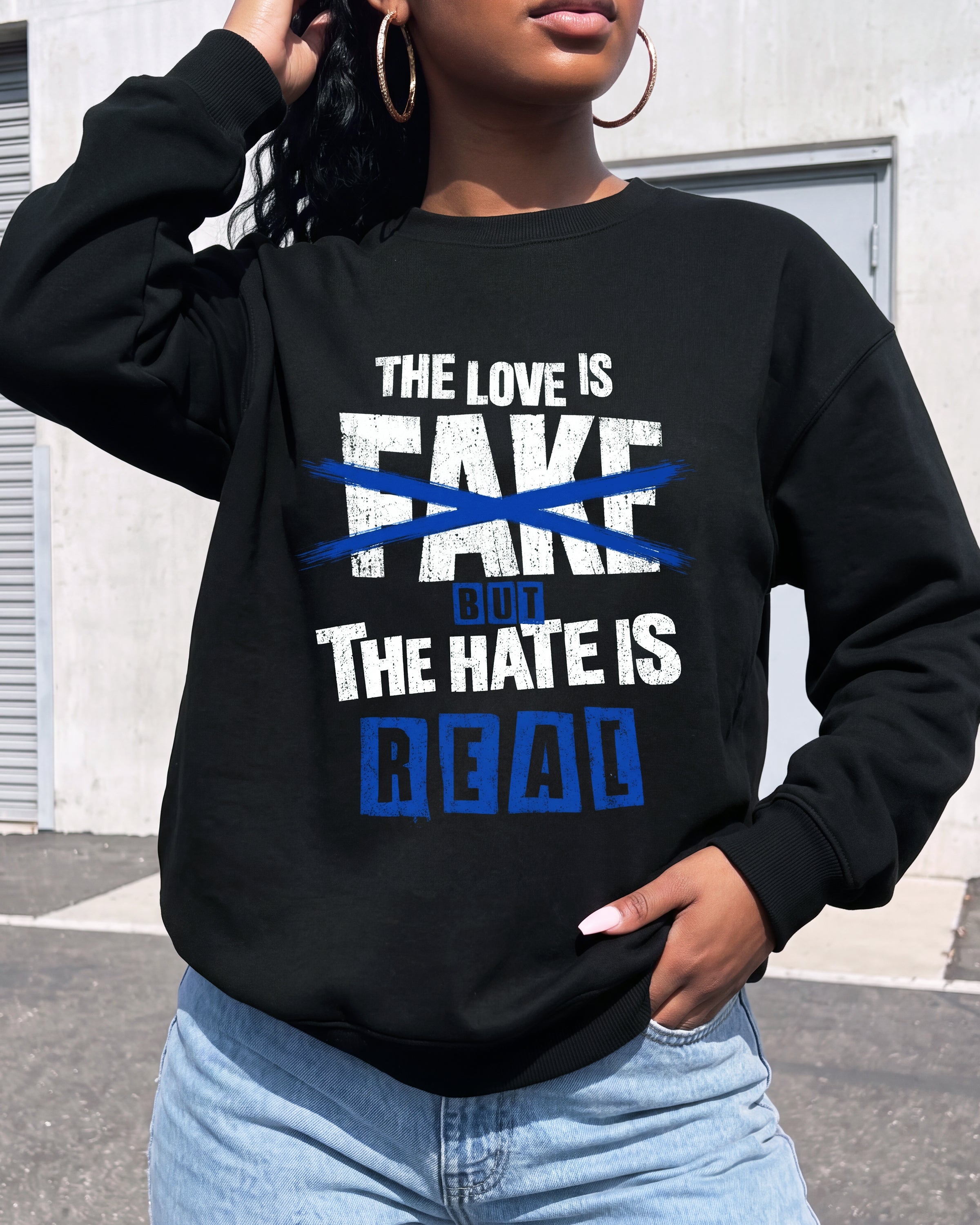A woman wearing a black crewneck sweatshirt with a graphic print that reads 'THE LOVE IS FAKE' crossed out in blue, and 'BUT THE HATE IS REAL' in blue block letters. She is wearing light blue jeans and large hoop earrings, standing outdoors in front of a gray building.