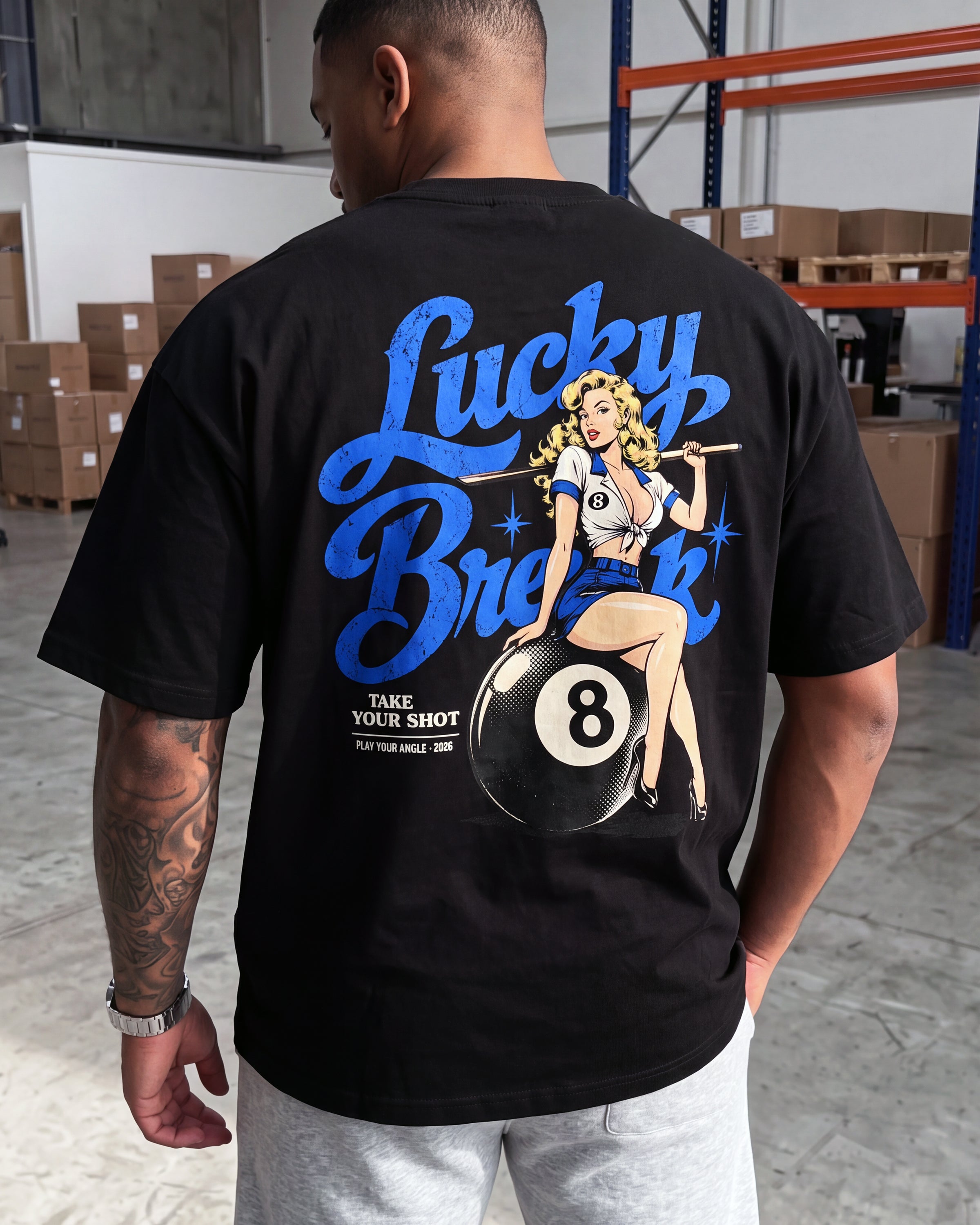 A man wearing a black graphic t-shirt with a large blue and white 'Lucky Break' pin-up girl and 8-ball design on the back, standing in a warehouse setting.