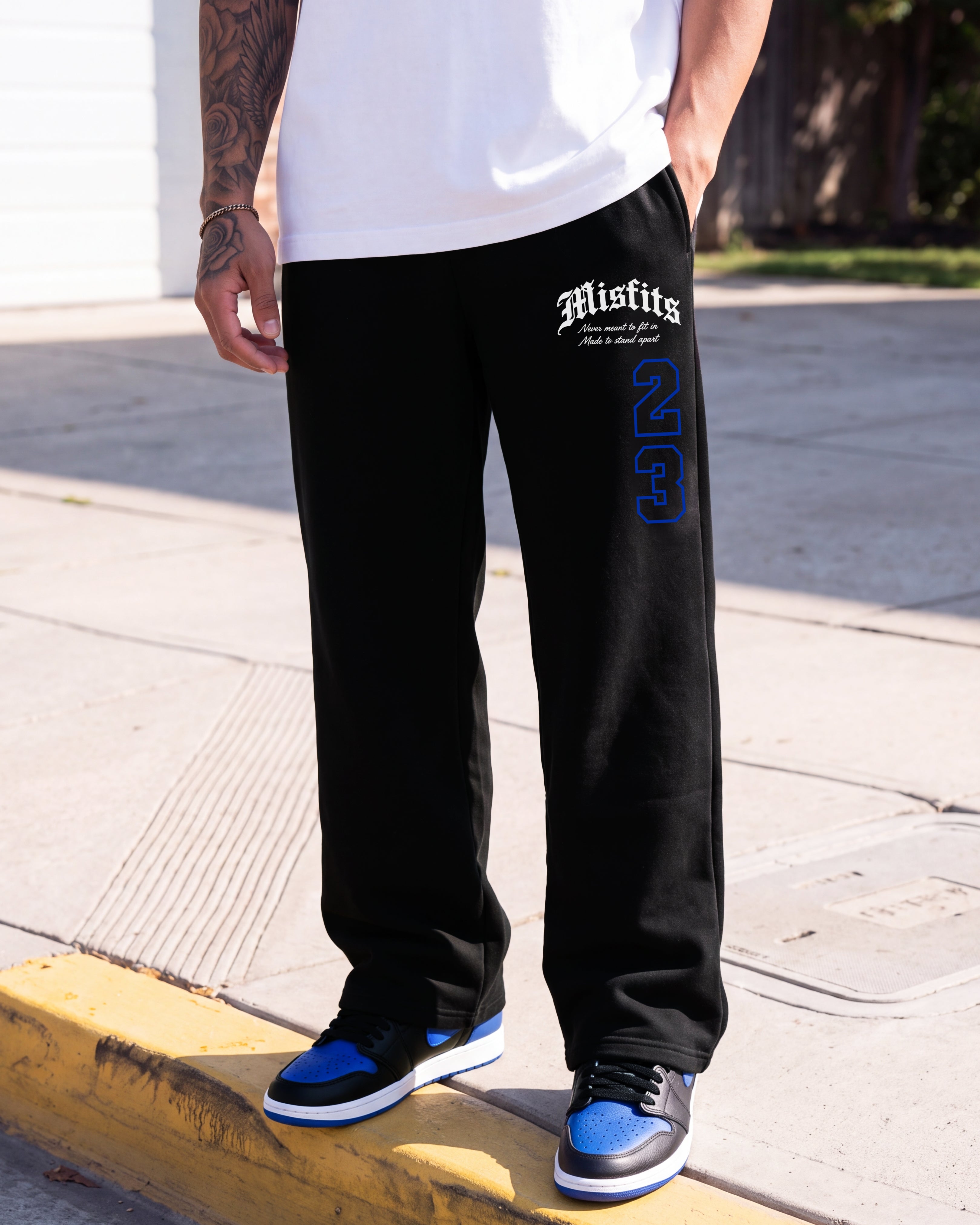 A person wearing black sweatpants with white and blue graphics, standing outdoors on a yellow curb. The sweatpants feature the word 'Misfits' in white gothic font and a large blue '26' outlined in white on the left leg.