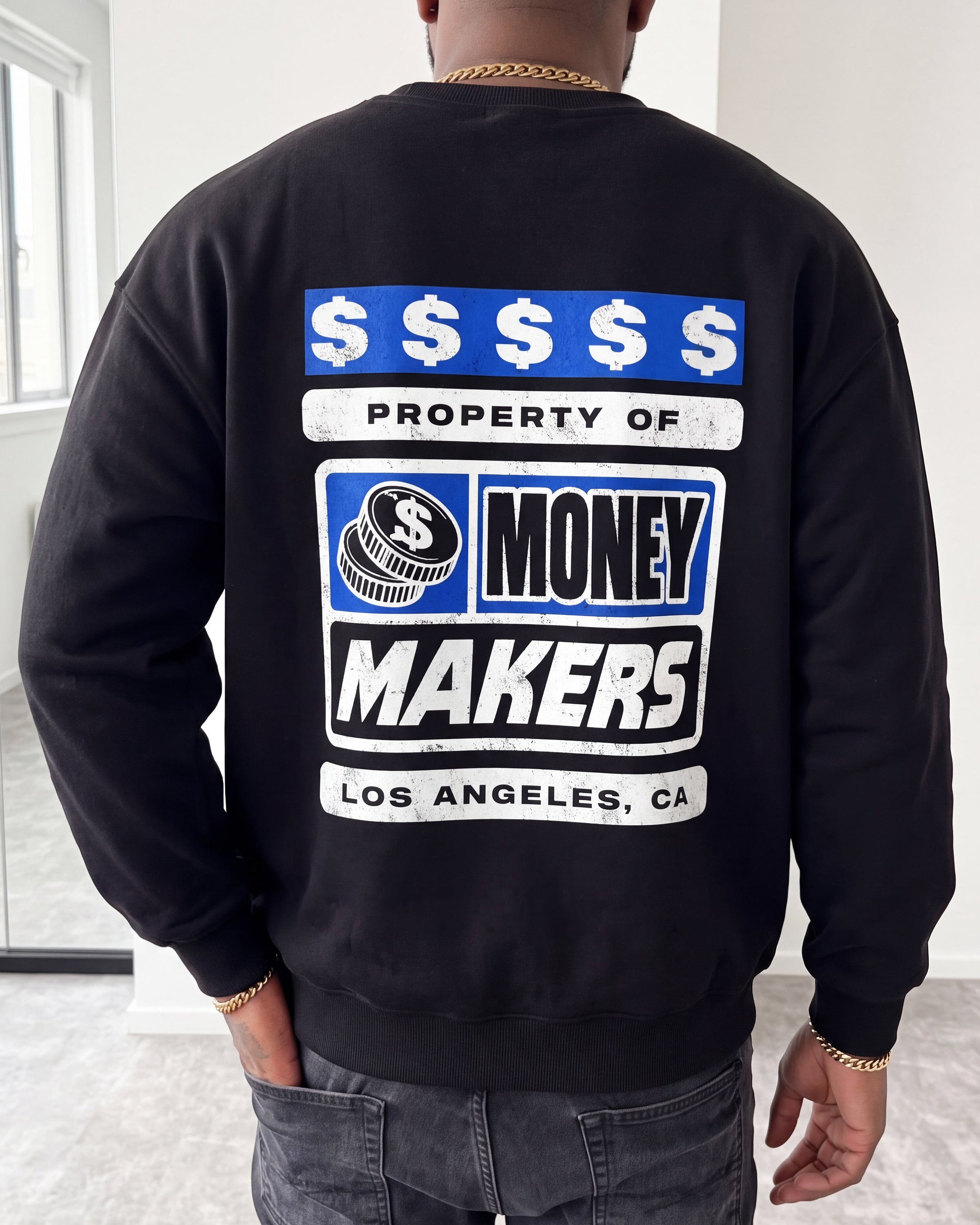 A person viewed from behind wearing a black crewneck sweatshirt with a large graphic print on the back. The graphic features blue and white dollar sign banners, a stack of coins with a dollar sign, and the text 'PROPERTY OF MONEY MAKERS LOS ANGELES, CA'.