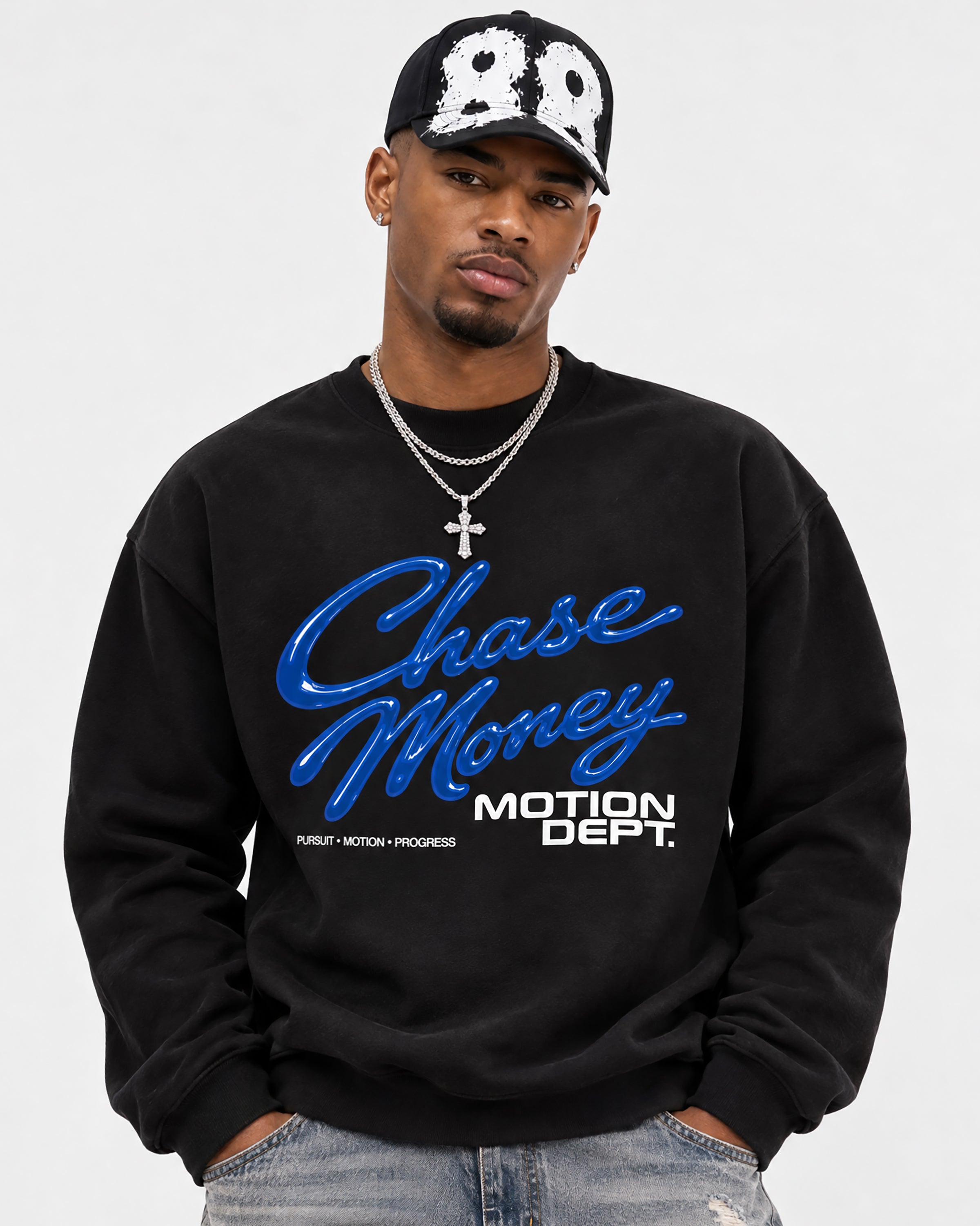 A man wearing a black crewneck sweatshirt with a blue and white graphic that reads 'Chase Money' and 'MOTION DEPT.', paired with a black baseball cap and jeans, against a light gray background.