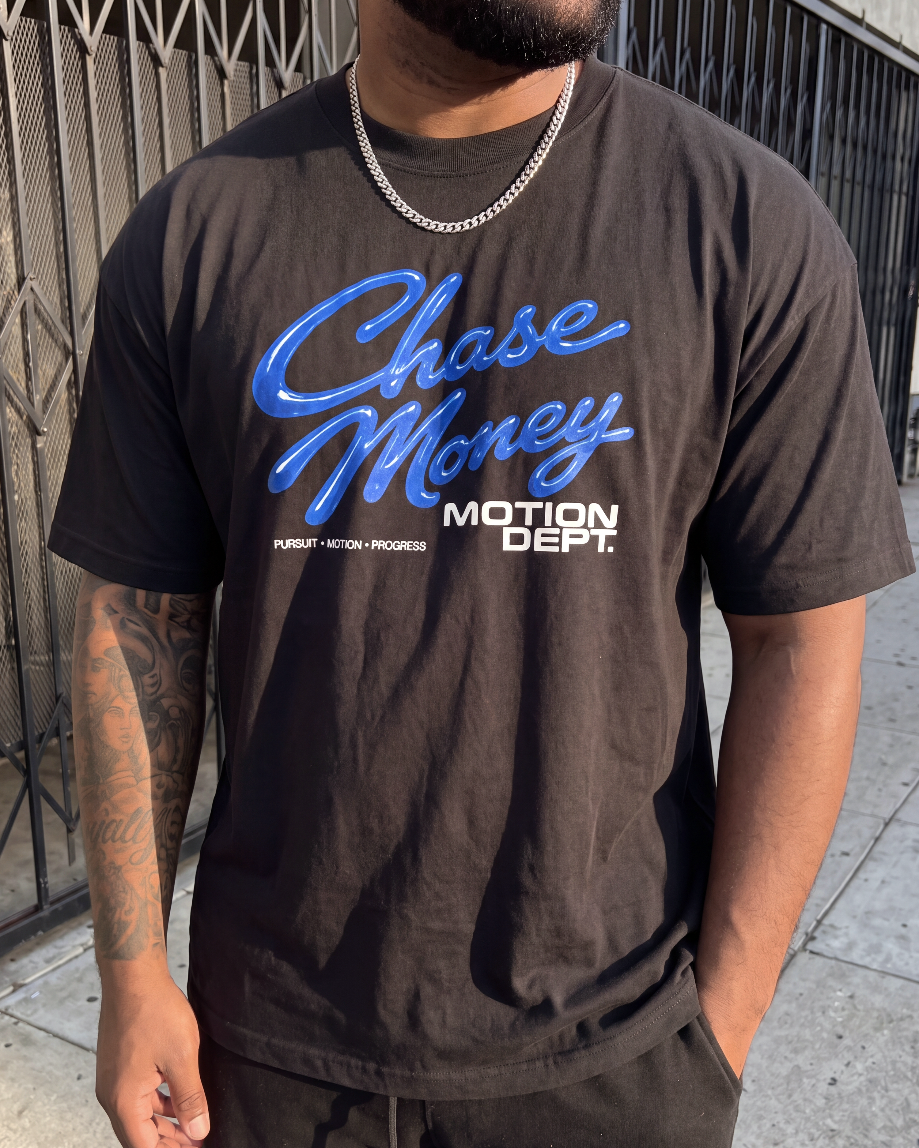 A man wearing a dark brown t-shirt with a blue and white graphic that reads 'Chase Money' and 'MOTION DEPT.' on the front, standing outdoors in front of a metal gate.