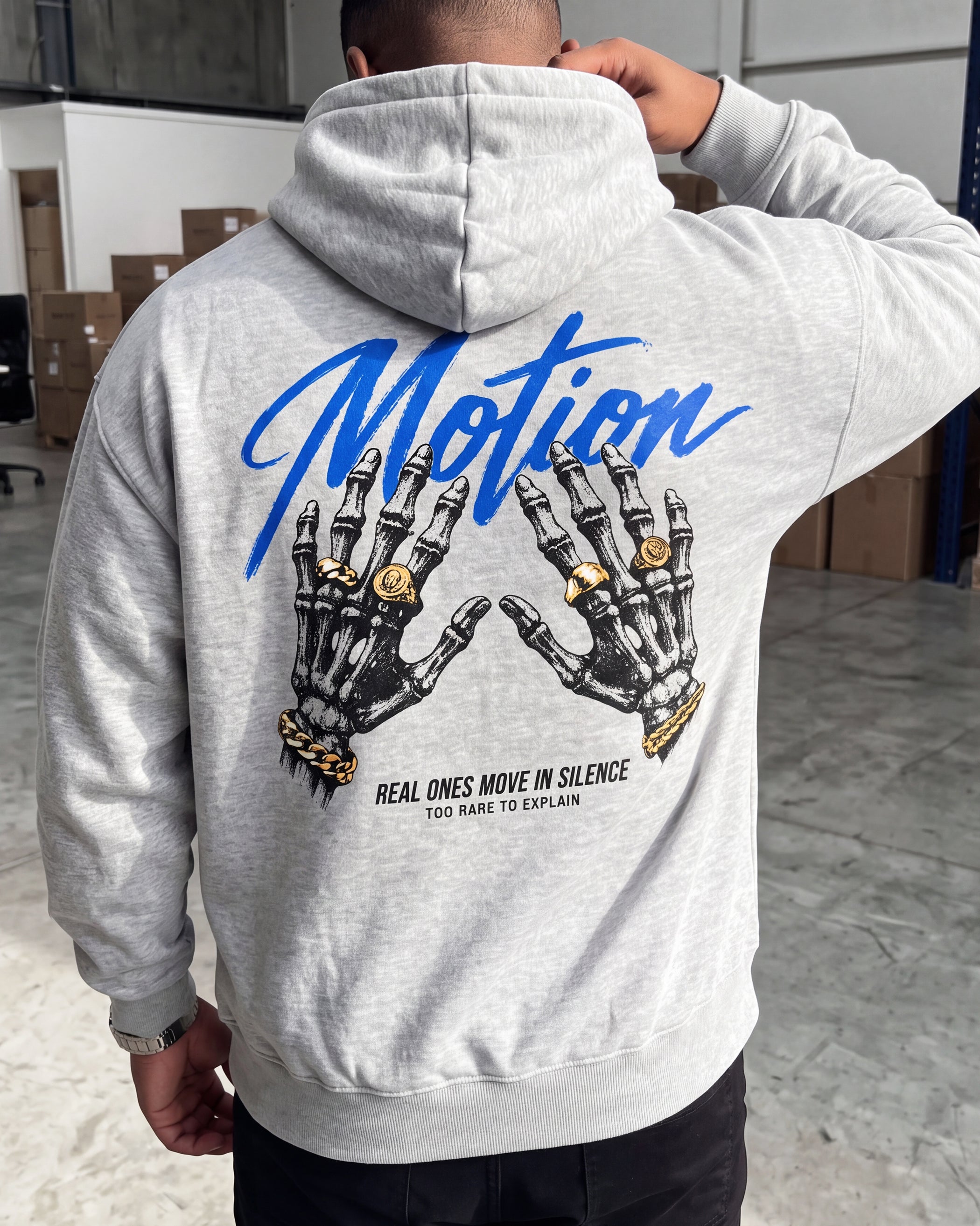 A person shown from the back wearing a light heather gray hoodie with a large graphic print on the back. The print features the word 'Motion' in blue script, two black-and-white skeletal hands wearing gold rings, and small black text reading 'REAL ONES MOVE IN SILENCE TOO RARE TO EXPLAIN.