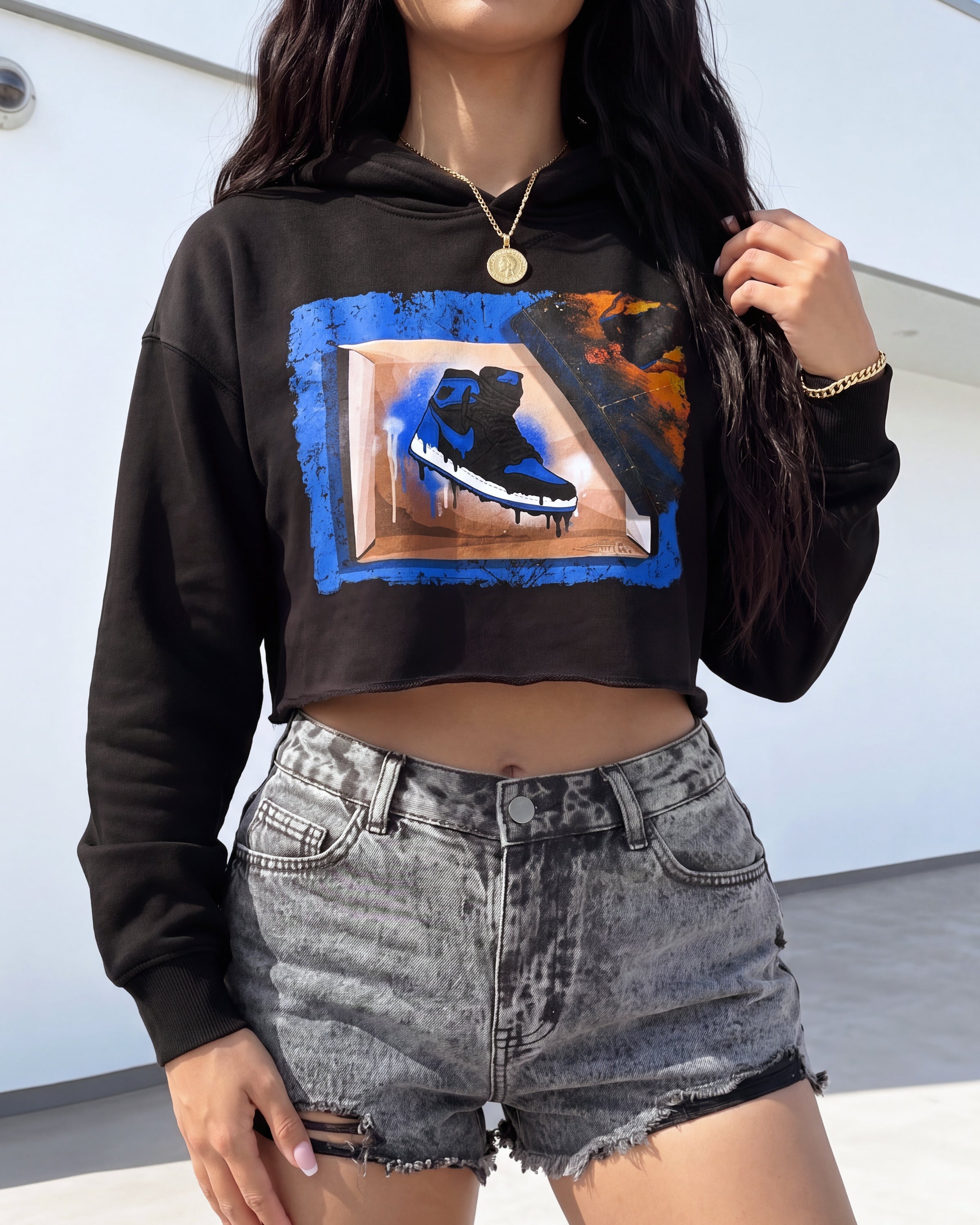 A woman wearing a black cropped hoodie with a large graphic print of a blue sneaker in a box, paired with gray acid-wash denim shorts.