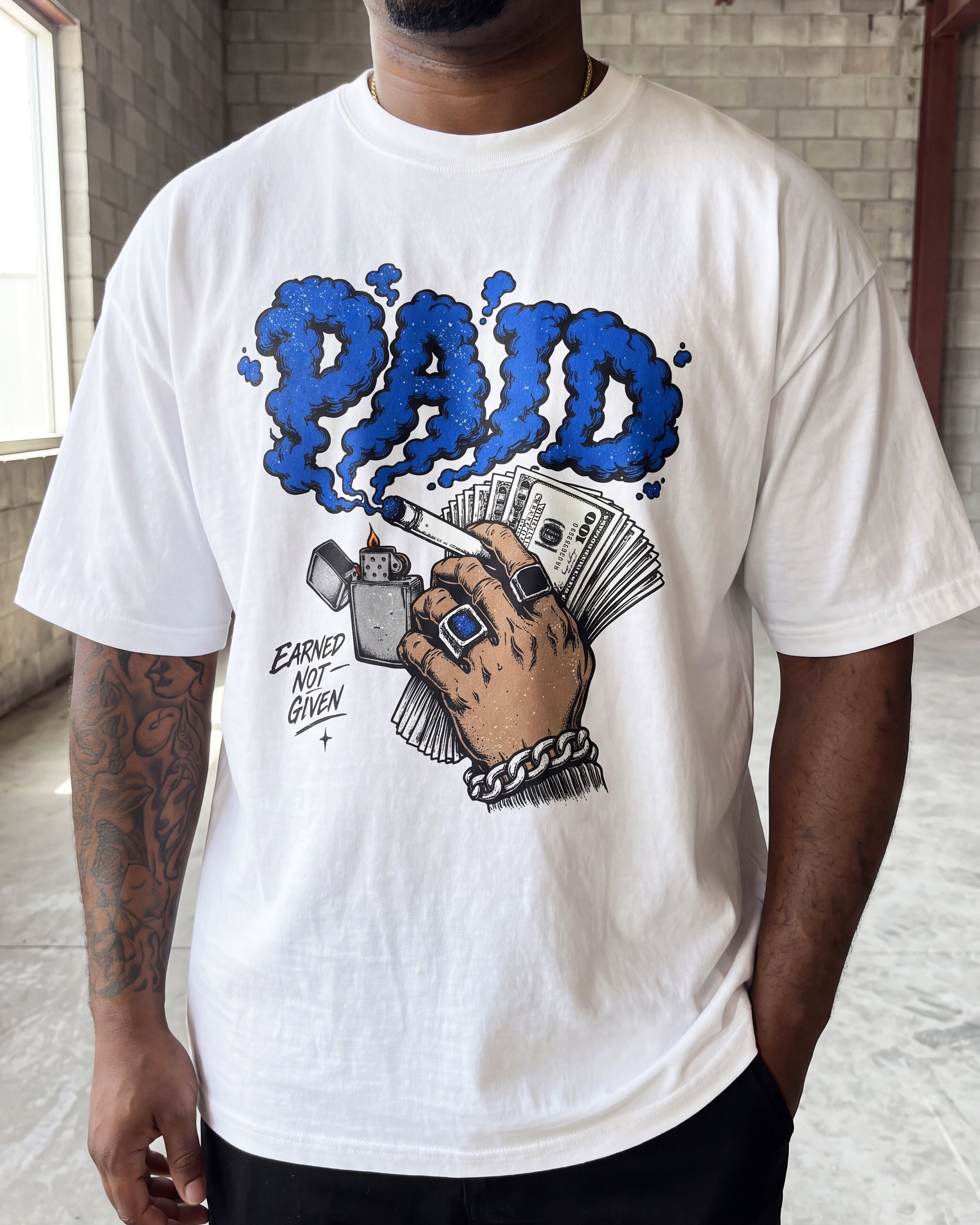A man wearing a white graphic t-shirt with a blue and black 'PAID' design on the front, standing in a brick hallway.