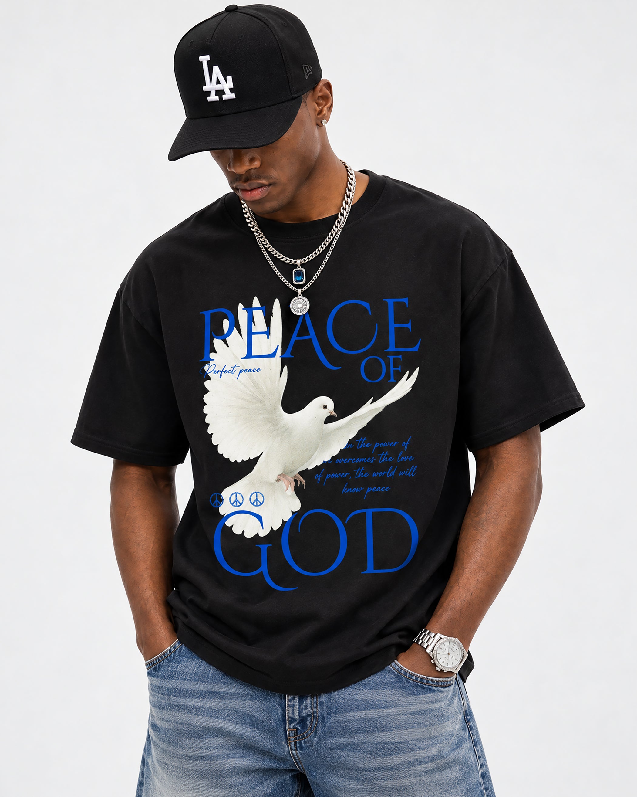 A man wearing a black graphic t-shirt with a white dove and blue 'PEACE OF GOD' text, paired with a black LA baseball cap, silver chains, and blue jeans, against a light gray background.