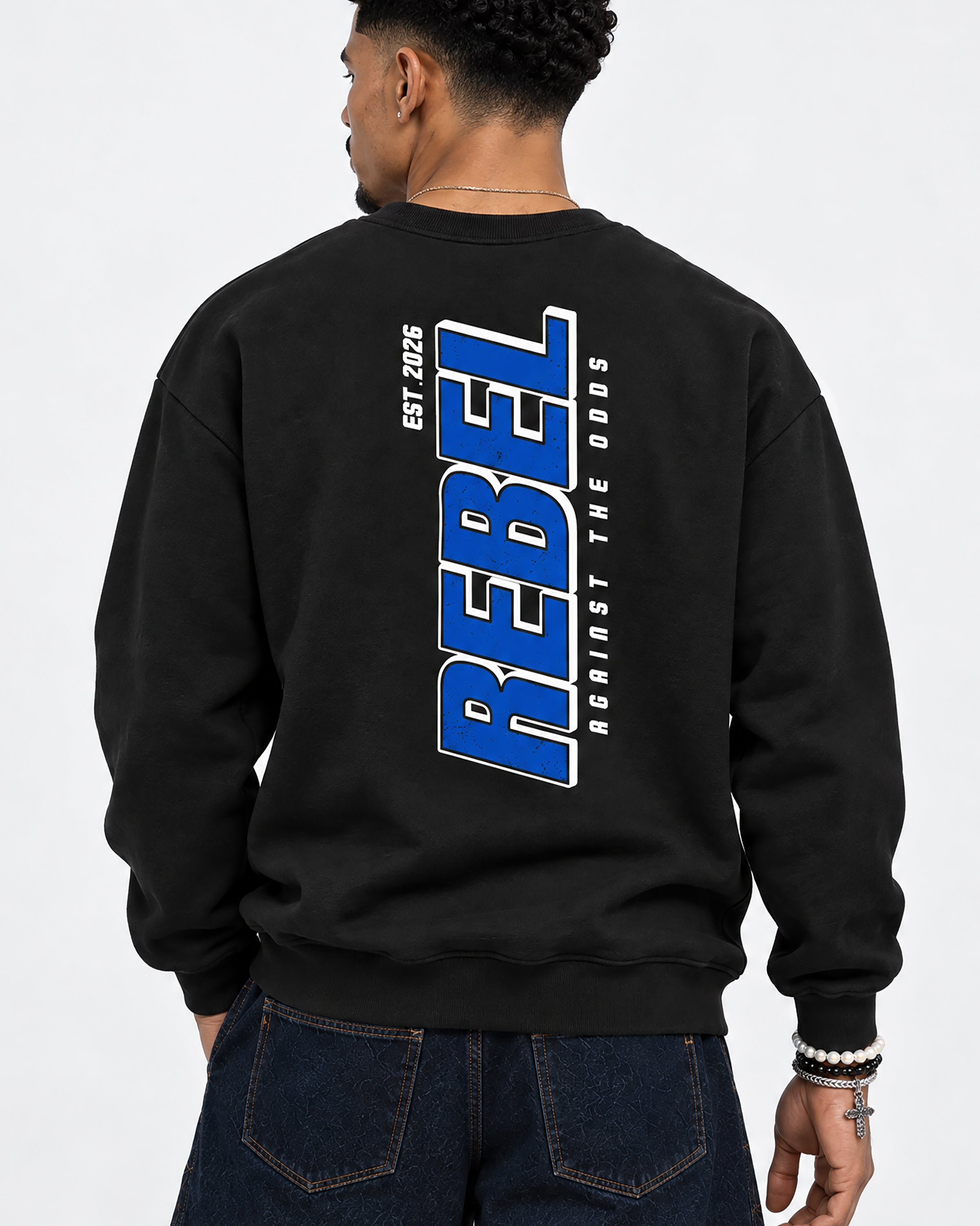 A man wearing a black crewneck sweatshirt from the back, featuring a large blue and white graphic print on the back that reads 'REBEL EST. 2025 AGAINST THE ODDS'.