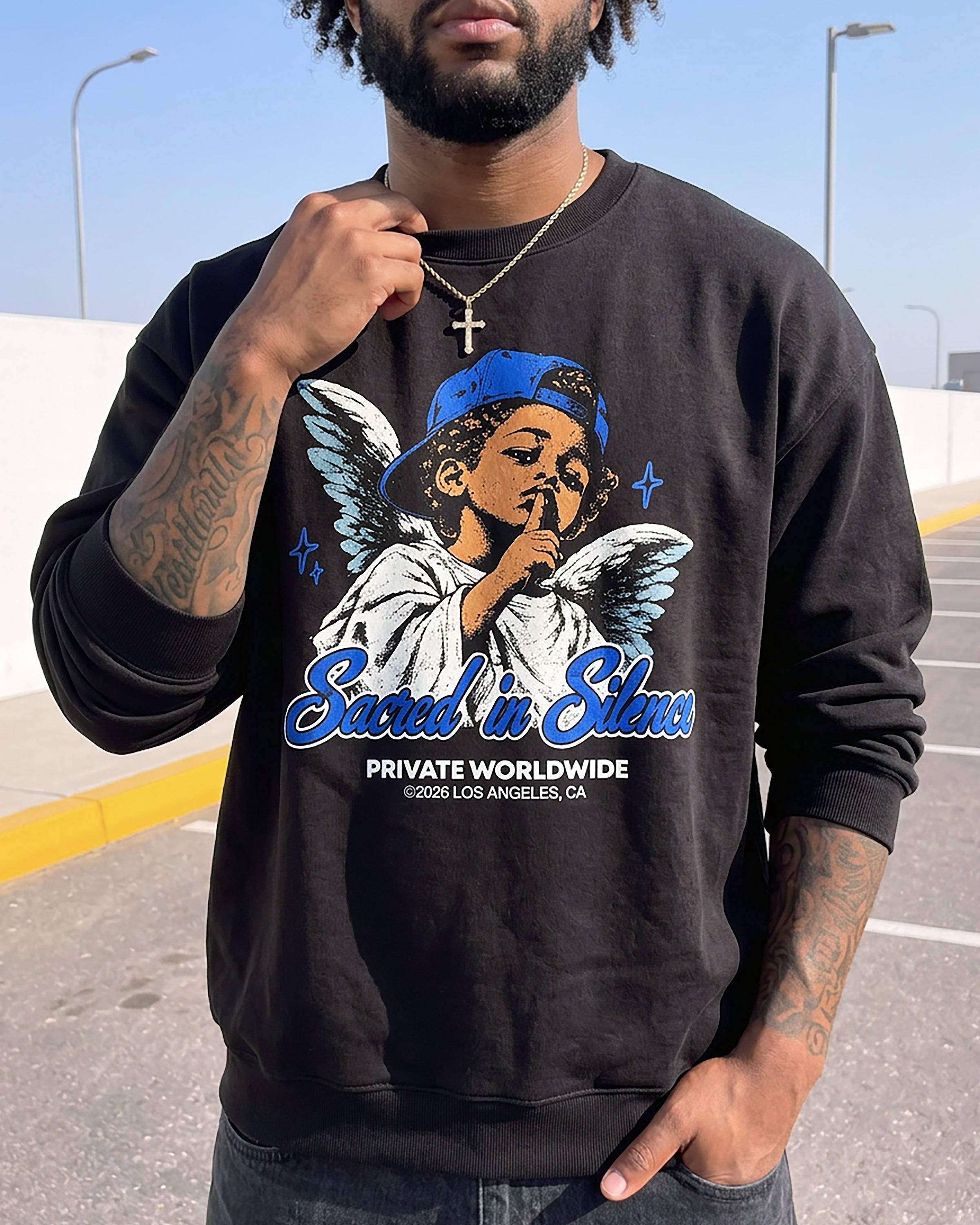 A man wearing a black crewneck sweatshirt with a colorful graphic of an angel and the text 'Sacred in Silence' and 'PRIVATE WORLDWIDE LOS ANGELES, CA'.