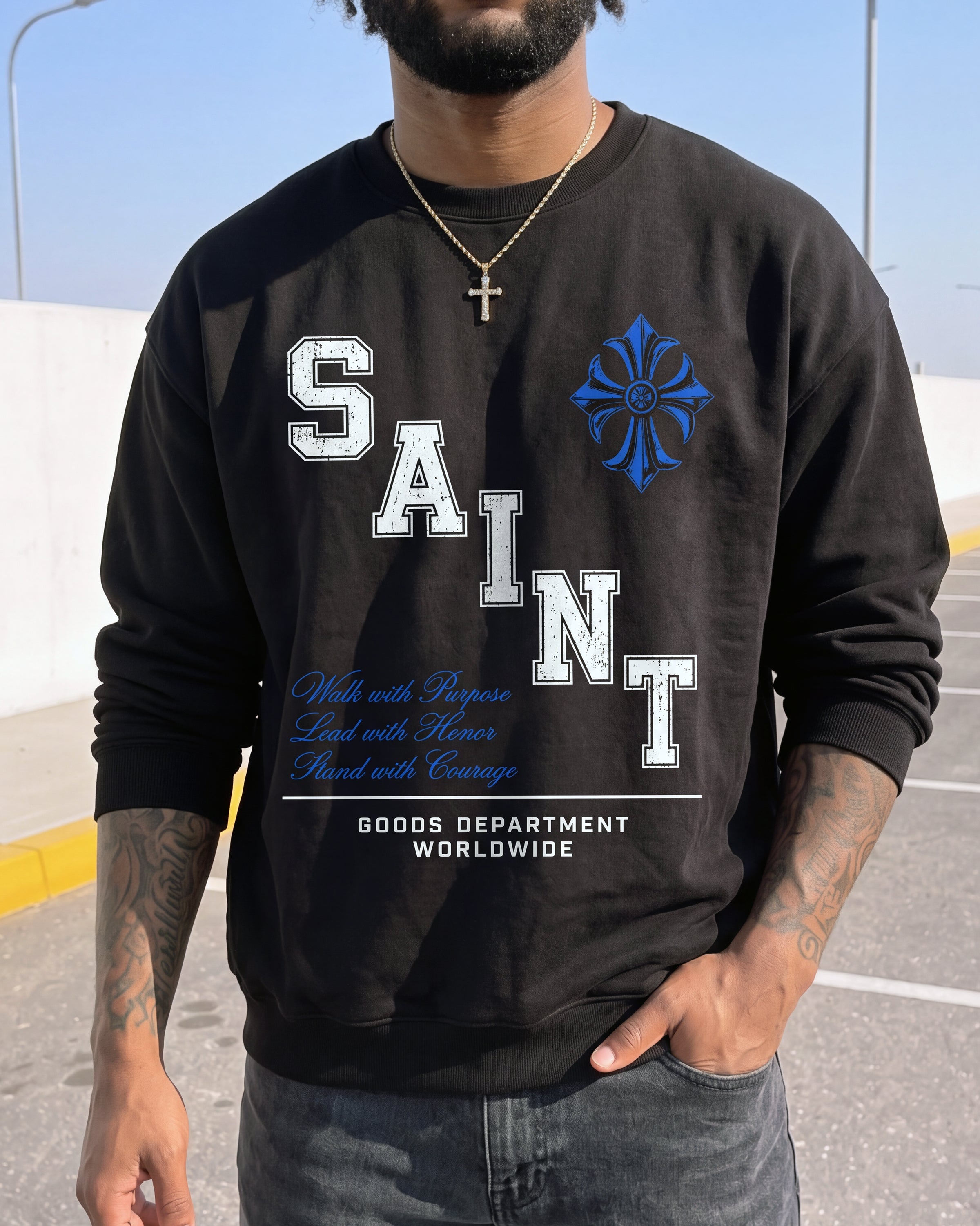 A man wearing a black crewneck sweatshirt with a graphic print. The sweatshirt features the word 'SAINT' in large white block letters with a blue cross design integrated into the lettering, along with blue and white text and 'GOODS DEPARTMENT WORLDWIDE' at the bottom.