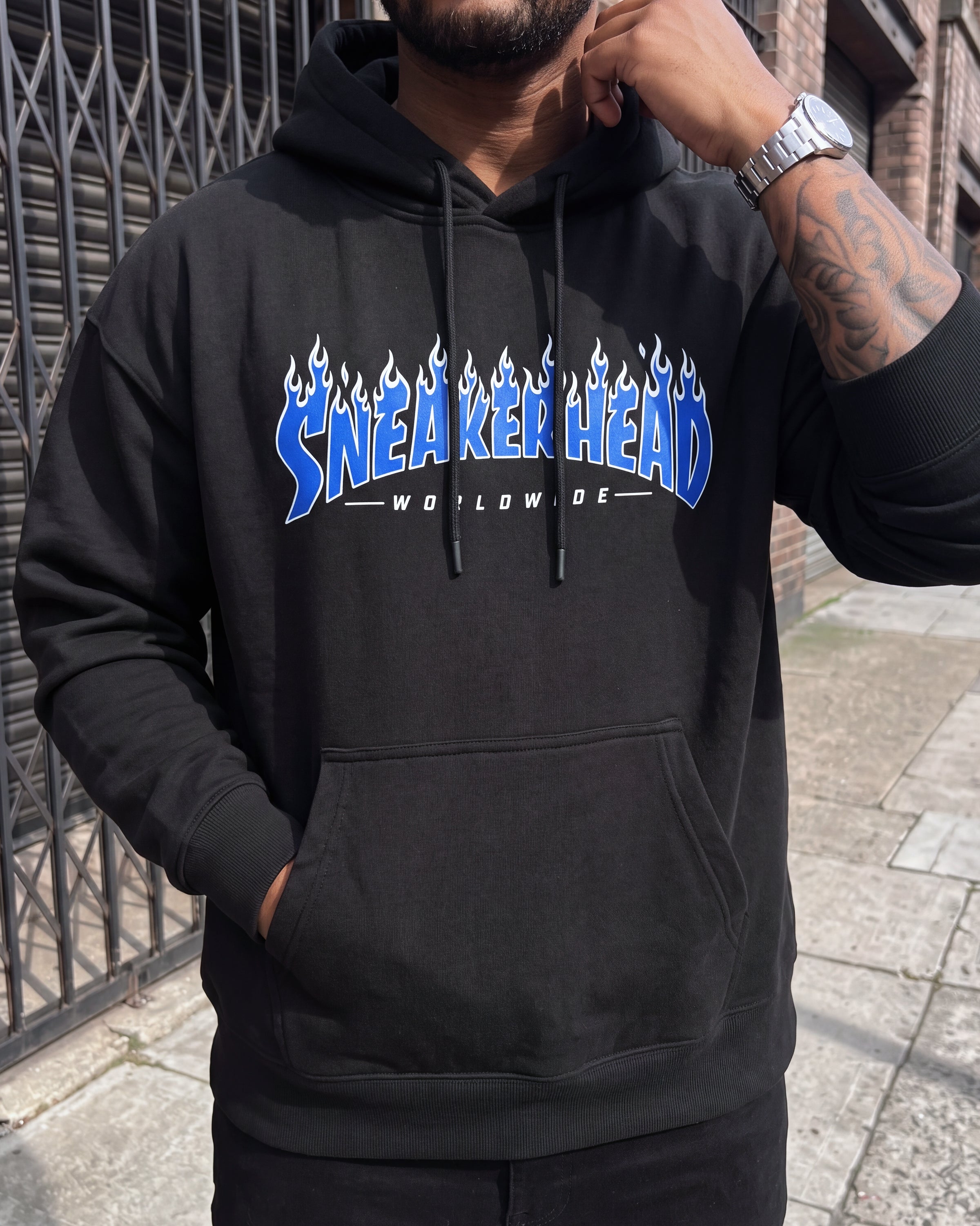 A man wearing a black pullover hoodie with a blue and white flame-style 'SNEAKERHEAD WORLDWIDE' graphic on the chest, standing outdoors near a metal gate and brick wall.