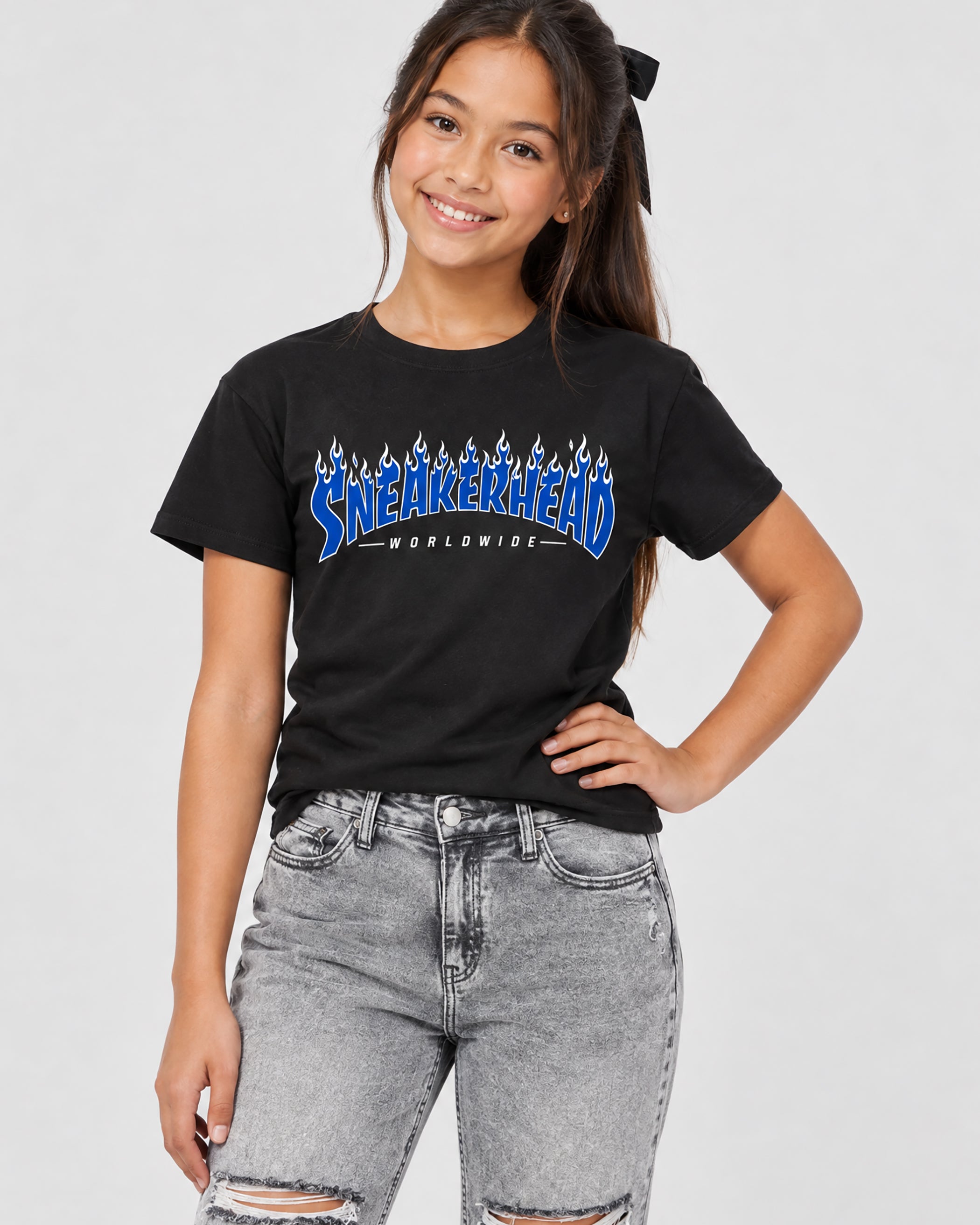 A young girl smiling and posing in a black crewneck t-shirt featuring a blue and white 'Sneakerhead Worldwide' graphic, paired with light-wash distressed jeans, against a solid light gray background.