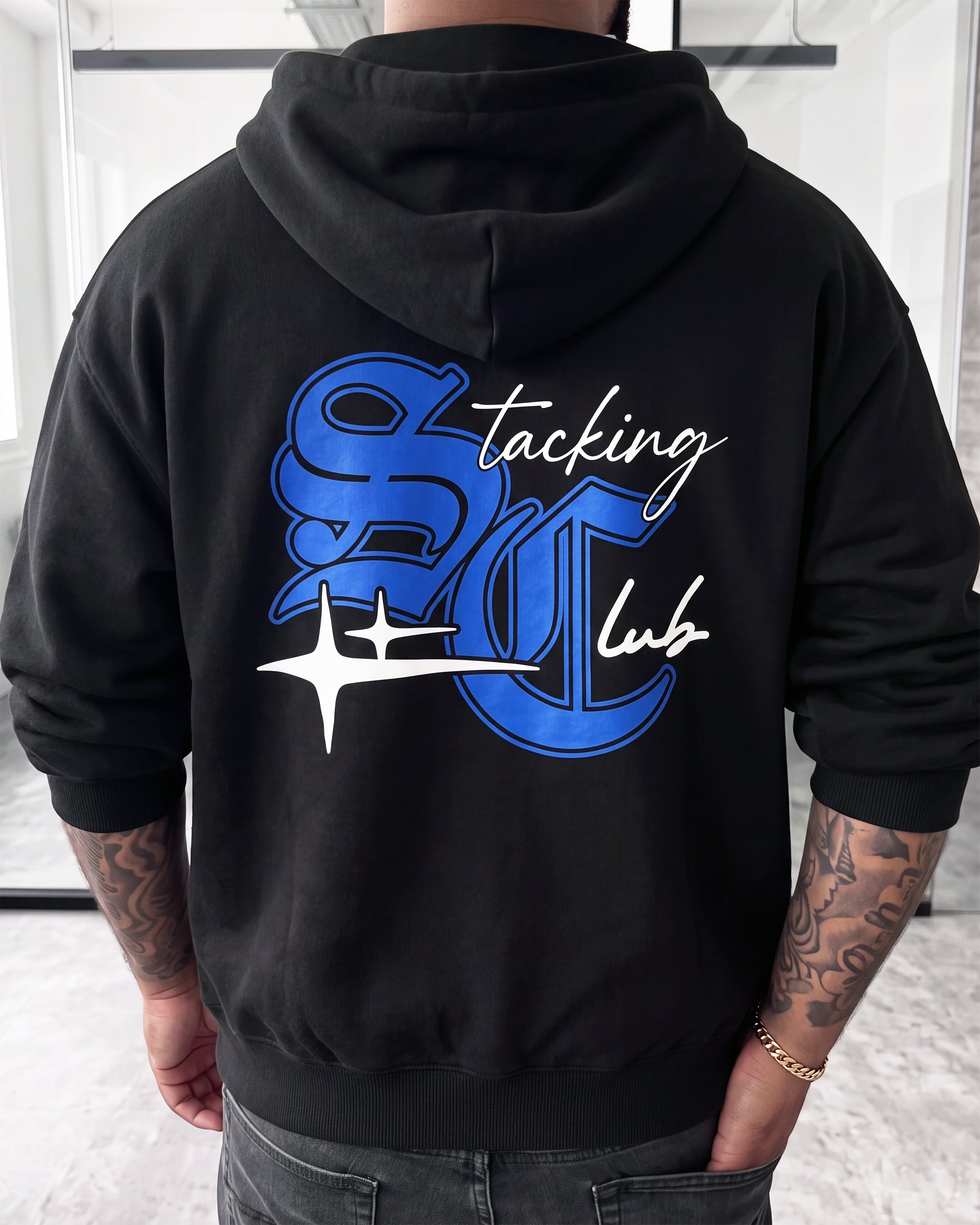A person shown from the back wearing a black hoodie with a large blue and white graphic on the back reading 'Stacking Club'.