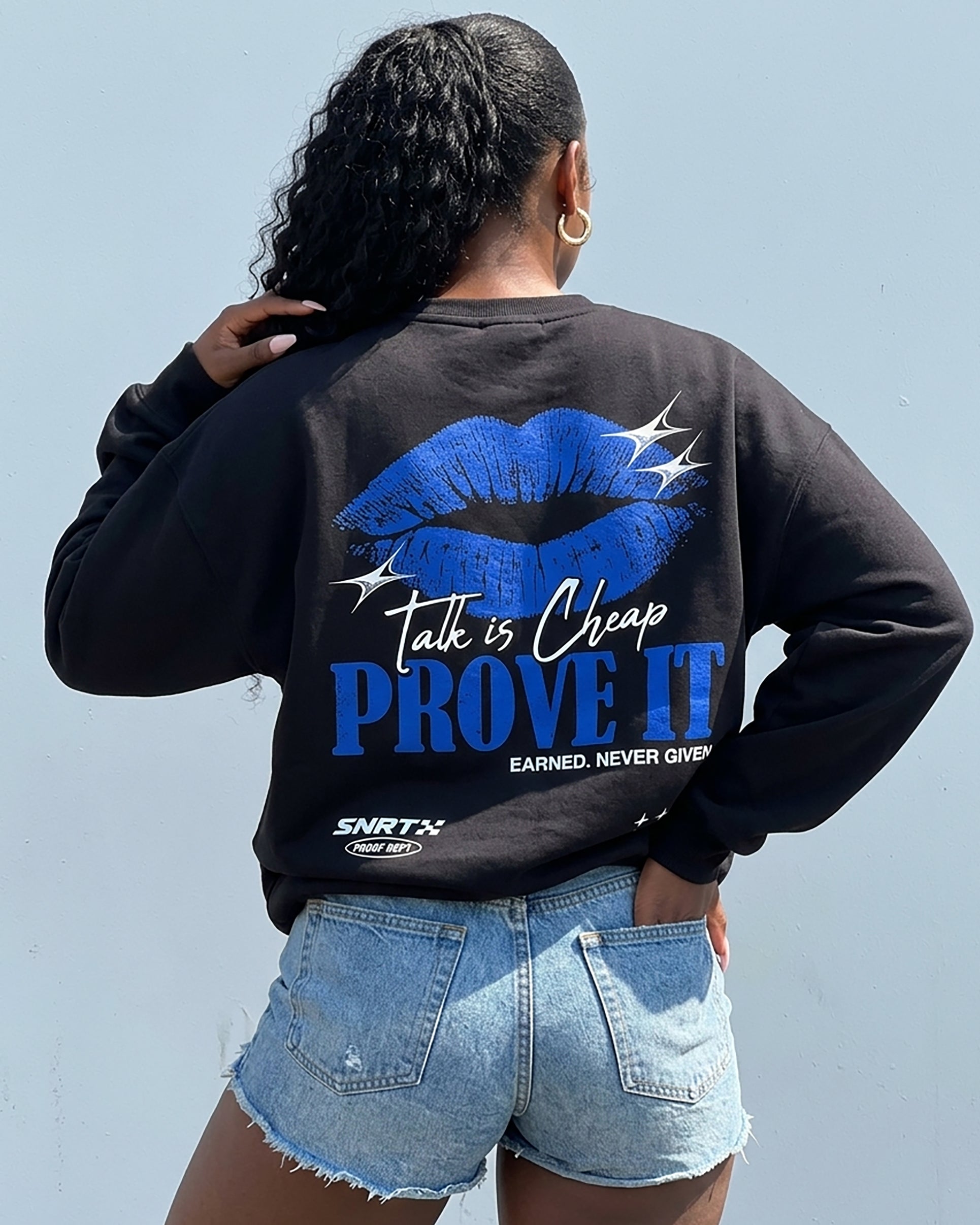 A woman shown from behind wearing a black sweatshirt with a large blue graphic on the back featuring lips and the text 'Talk is Cheap PROVE IT' and 'EARNED. NEVER GIVE UP.