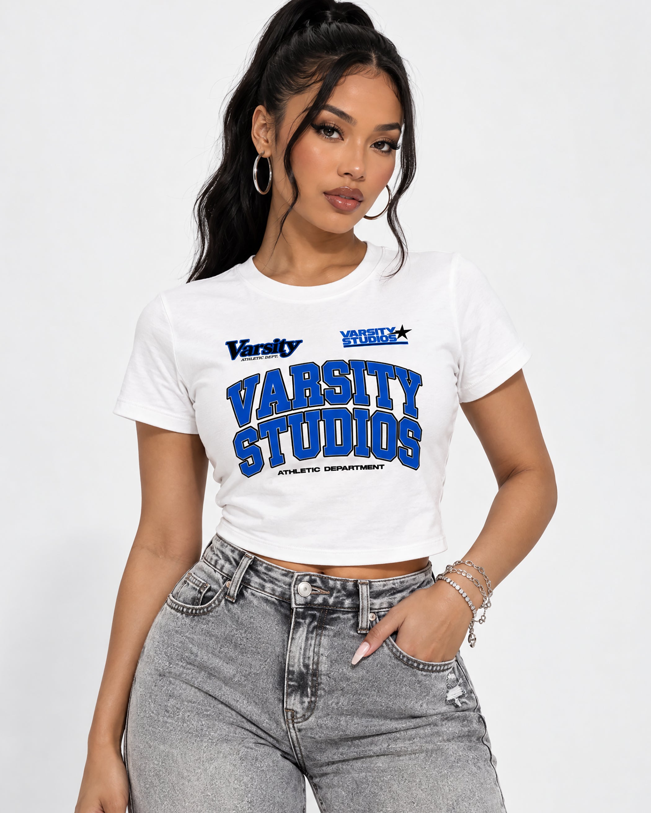 A woman modeling a white short-sleeve crop top with blue and navy varsity-style graphics reading 'Varsity Studios' and 'Athletic Department,' paired with gray jeans against a light gray studio background.