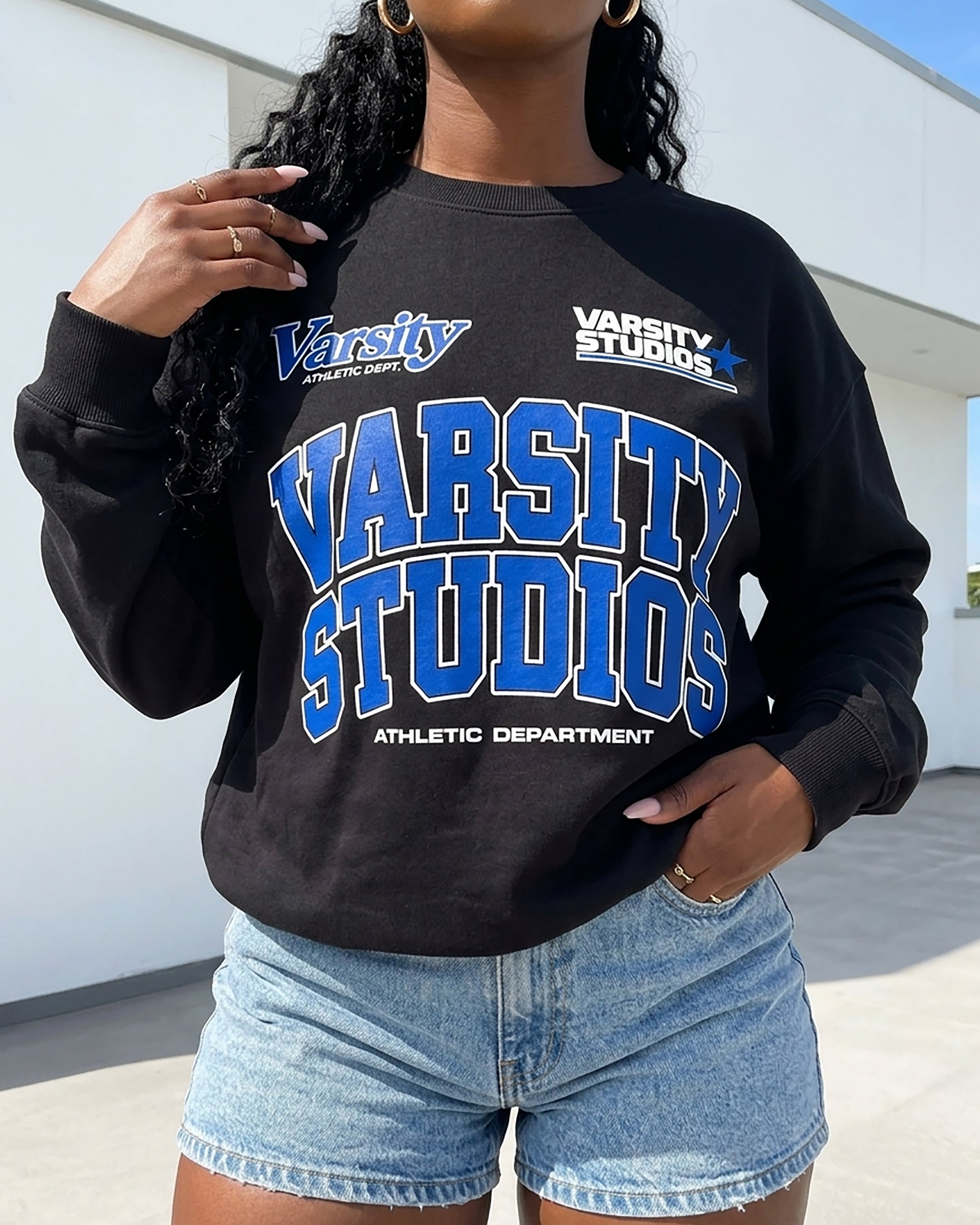A woman wearing a black long-sleeve sweatshirt with a blue and white 'Varsity Studios Athletic Department' graphic, paired with light-wash denim shorts, standing outdoors against a white building wall.