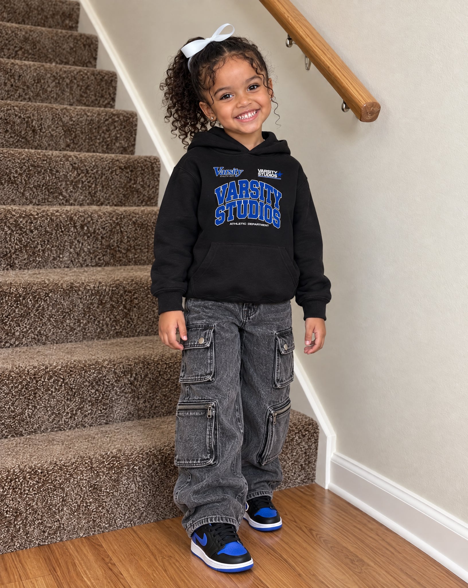 A young girl with curly hair stands on a carpeted staircase wearing a black hoodie with blue and white 'Varsity Studios' lettering, gray cargo pants, and blue and black sneakers.