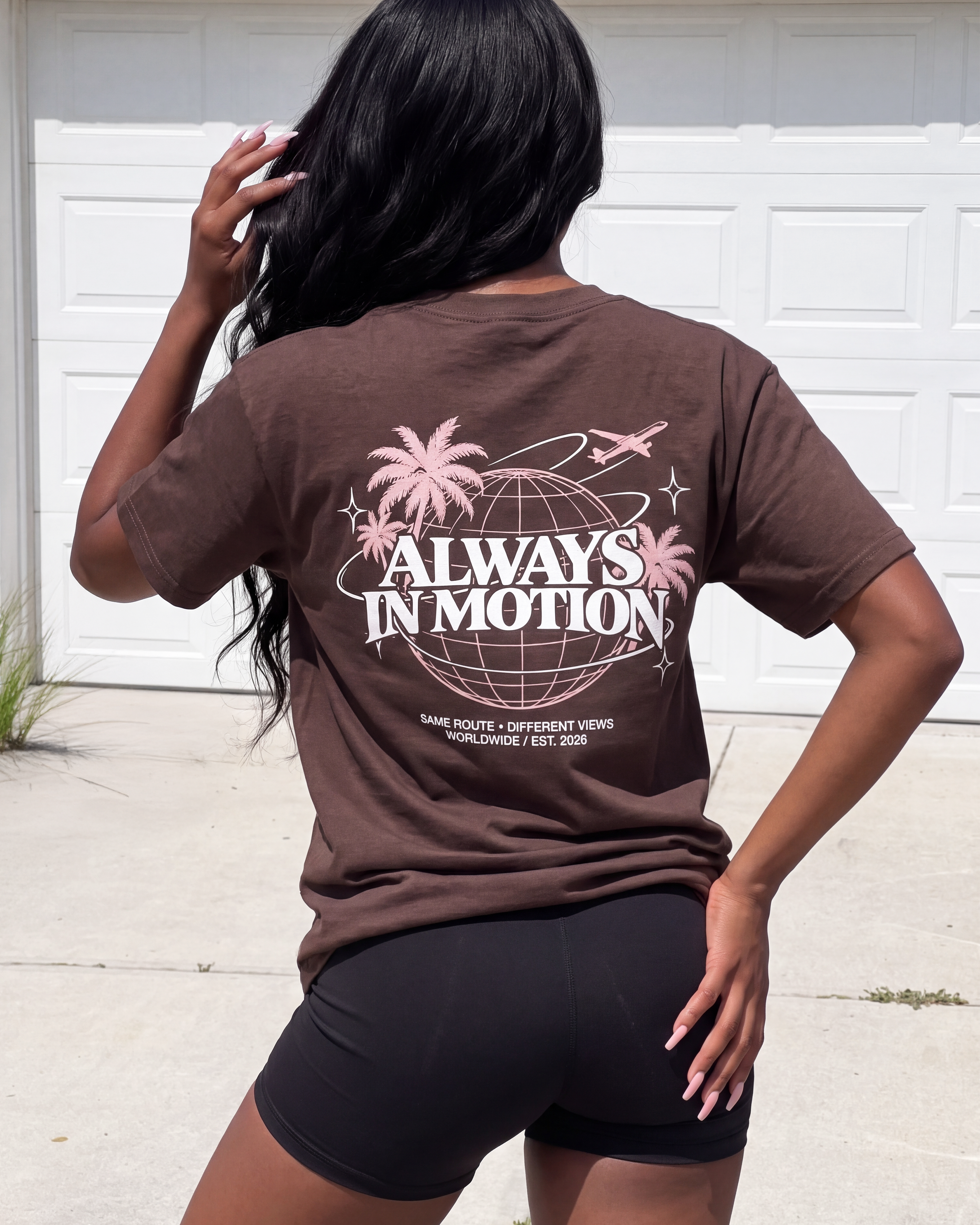 A woman wearing a brown graphic t-shirt and black bike shorts, photographed from behind in front of a white garage door. The back of the t-shirt features a pink and white graphic with the text 'ALWAYS IN MOTION' and a globe design.
