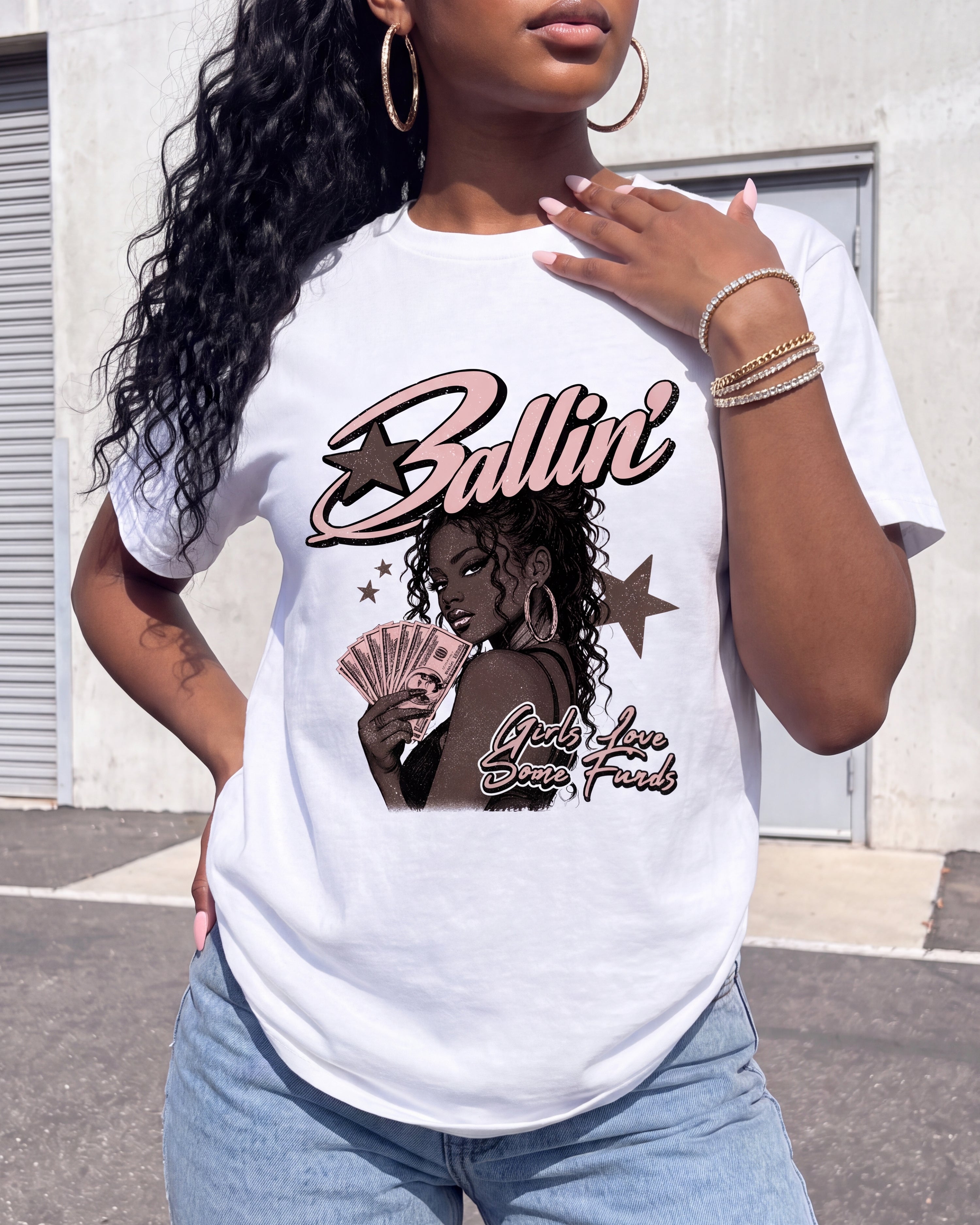 A woman wearing a white graphic t-shirt with a 'Ballin' Lady Love Cash Flow' design, paired with light blue jeans, standing outdoors in front of a building.