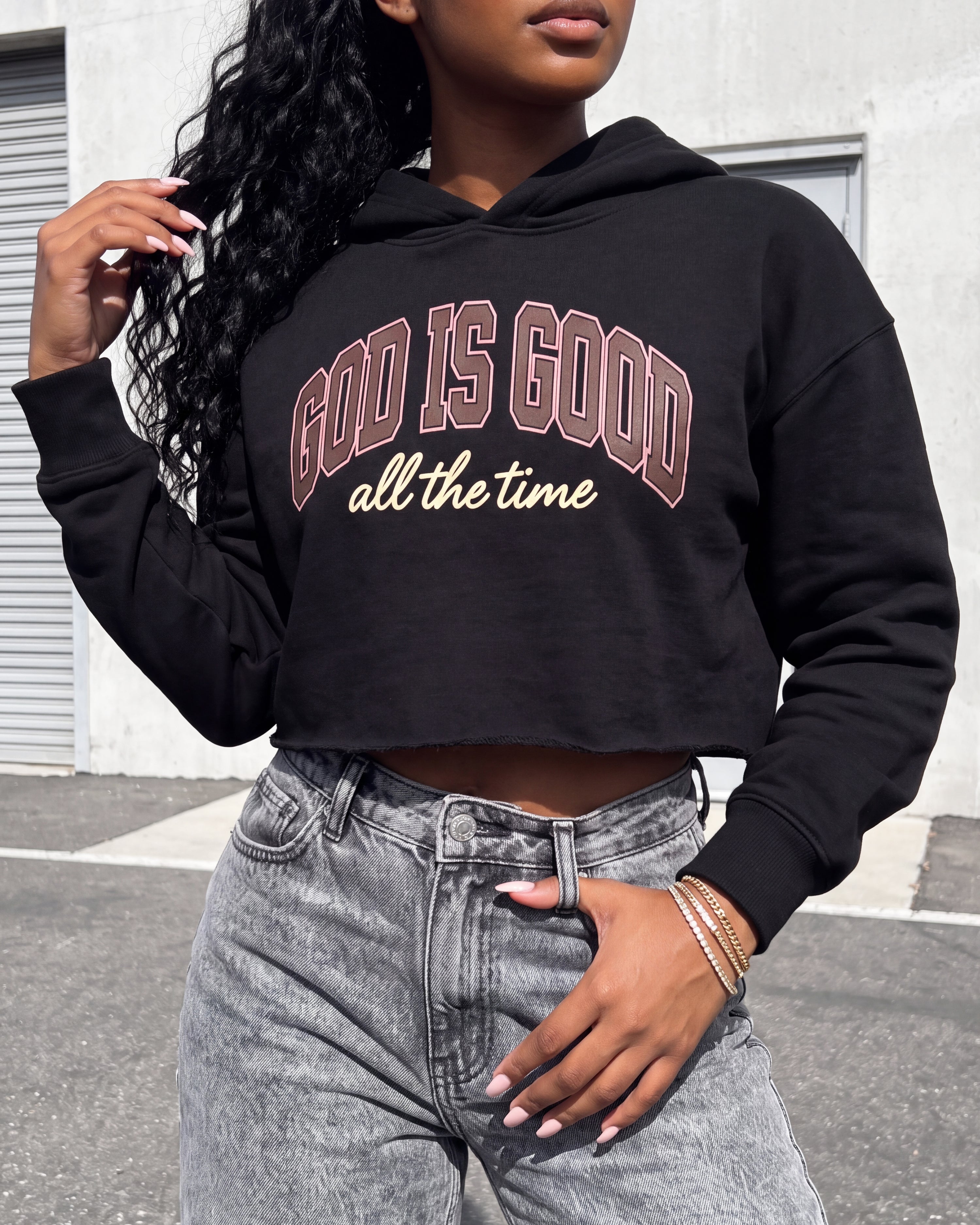 A woman wearing a black cropped hoodie with the text 'GOD IS GOOD all the time' printed on the front, paired with light-wash gray jeans, standing outdoors in front of a concrete wall.