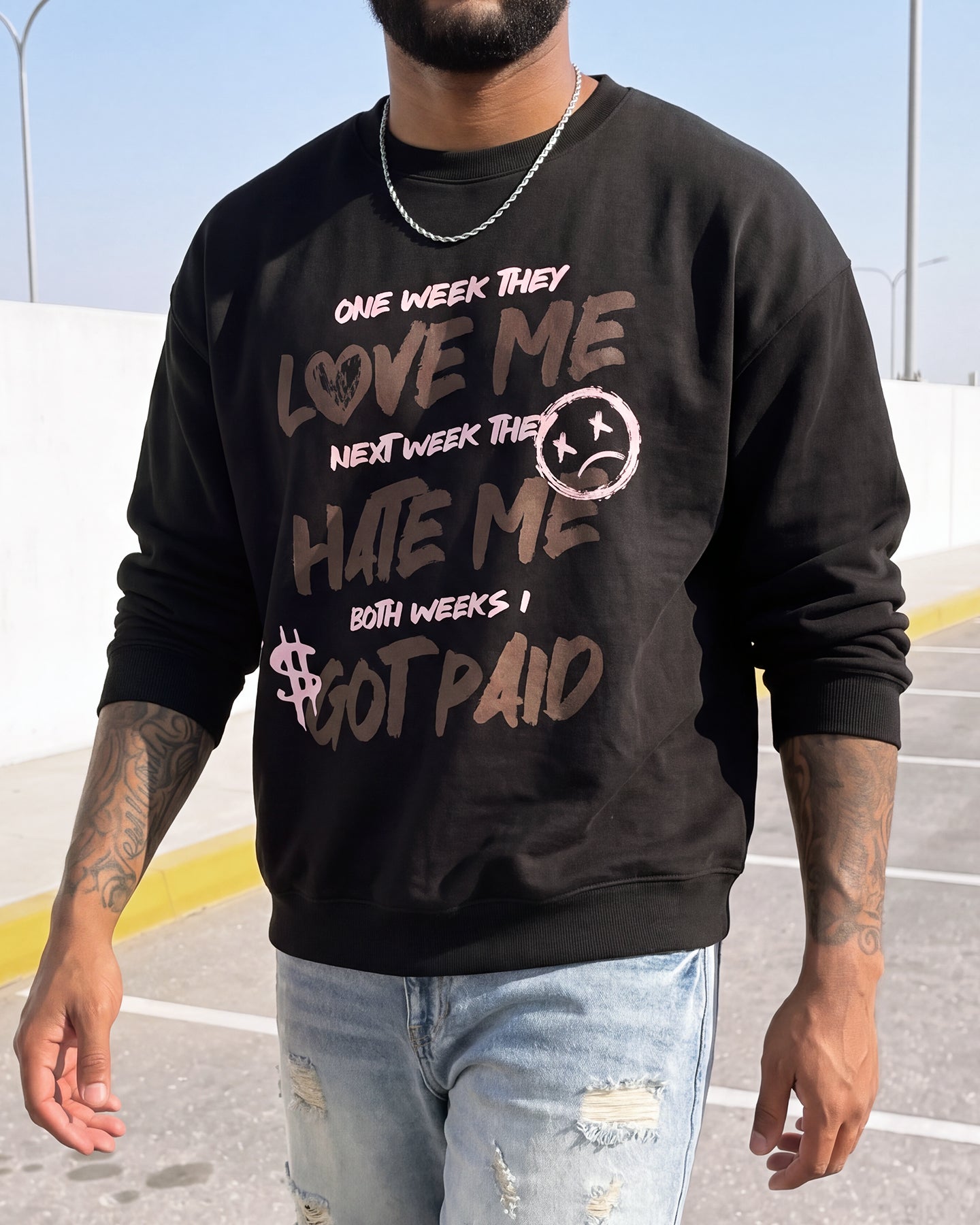 A man wearing a black crewneck sweatshirt with a distressed graphic that reads 'ONE WEEK THEY LOVE ME NEXT WEEK THEY HATE ME BOTH WEEKS I GOT PAID' in brown and pink lettering, paired with light blue ripped jeans.