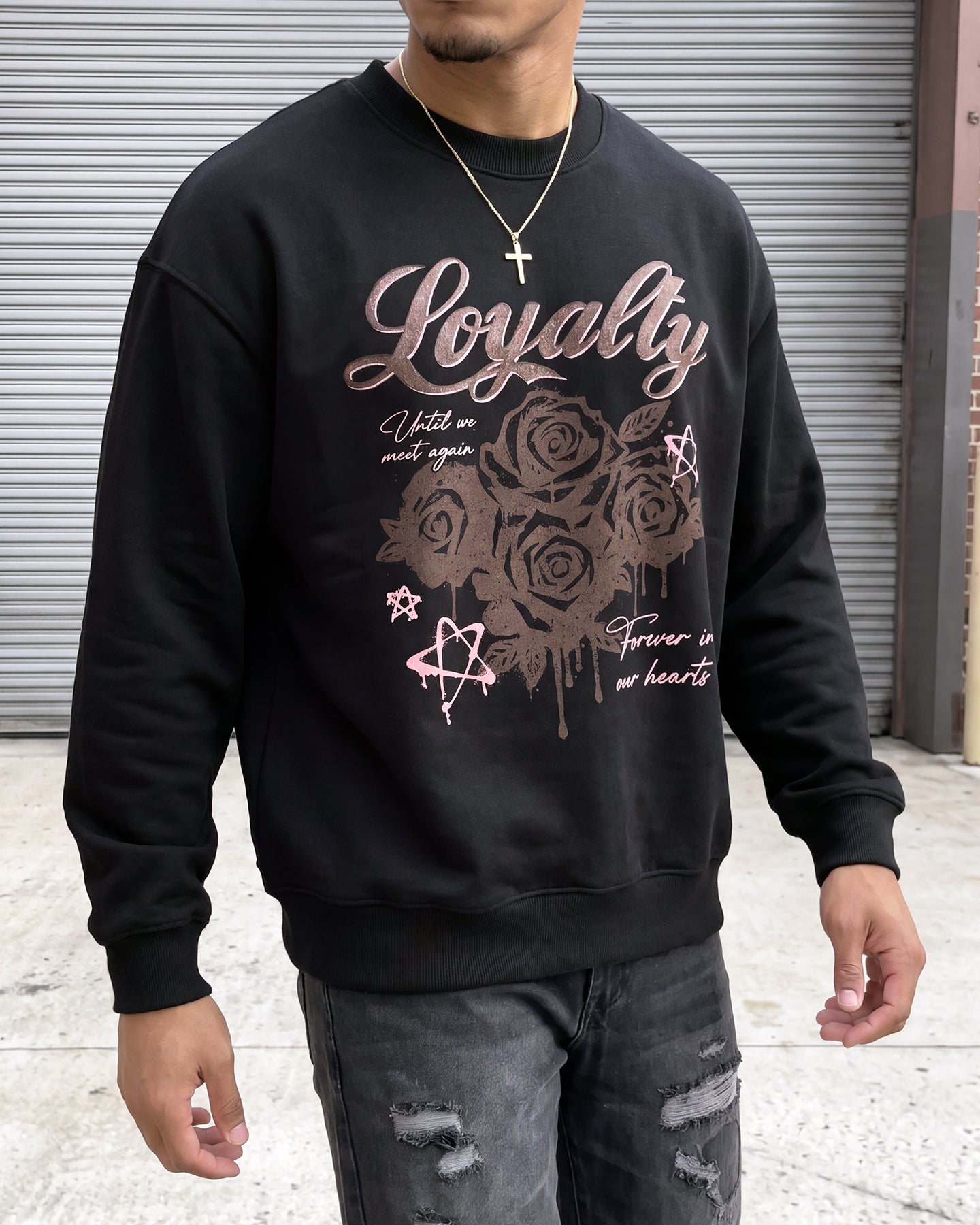A man wearing a black crewneck sweatshirt with a rose graphic and the word 'Loyalty' printed in rose gold on the front, paired with distressed gray jeans.