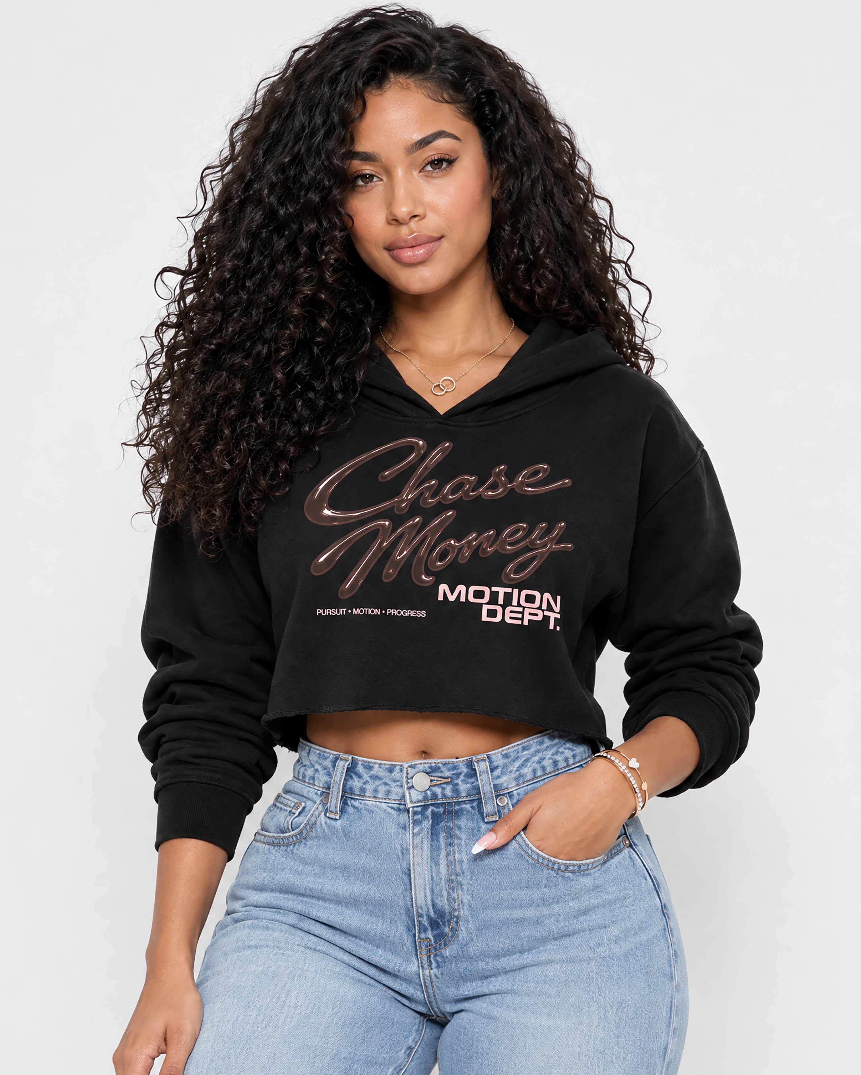 A woman with long, curly dark hair models a black cropped hoodie with 'Chase Money MOTION DEPT.' printed in metallic rose gold and white across the chest, paired with light blue jeans against a plain white studio background.
