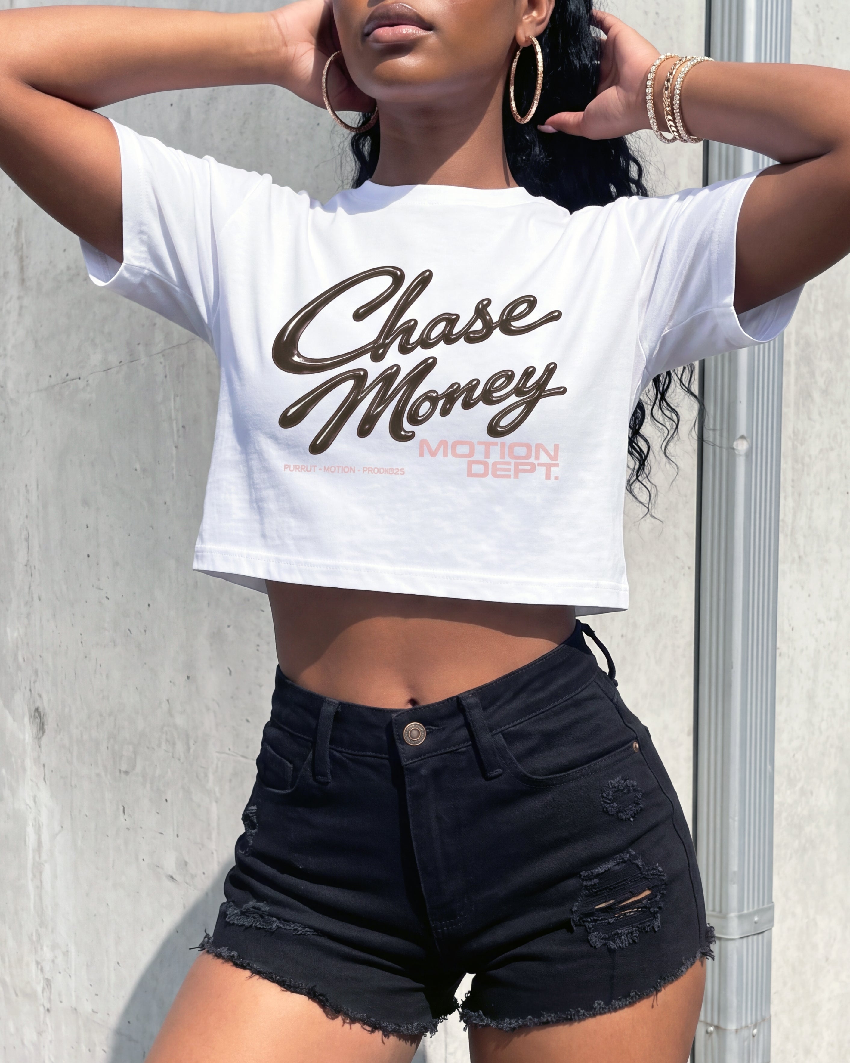 A woman wearing a white cropped graphic T-shirt with 'Chase Money' printed in brown script and distressed black denim shorts, posing against a concrete wall.