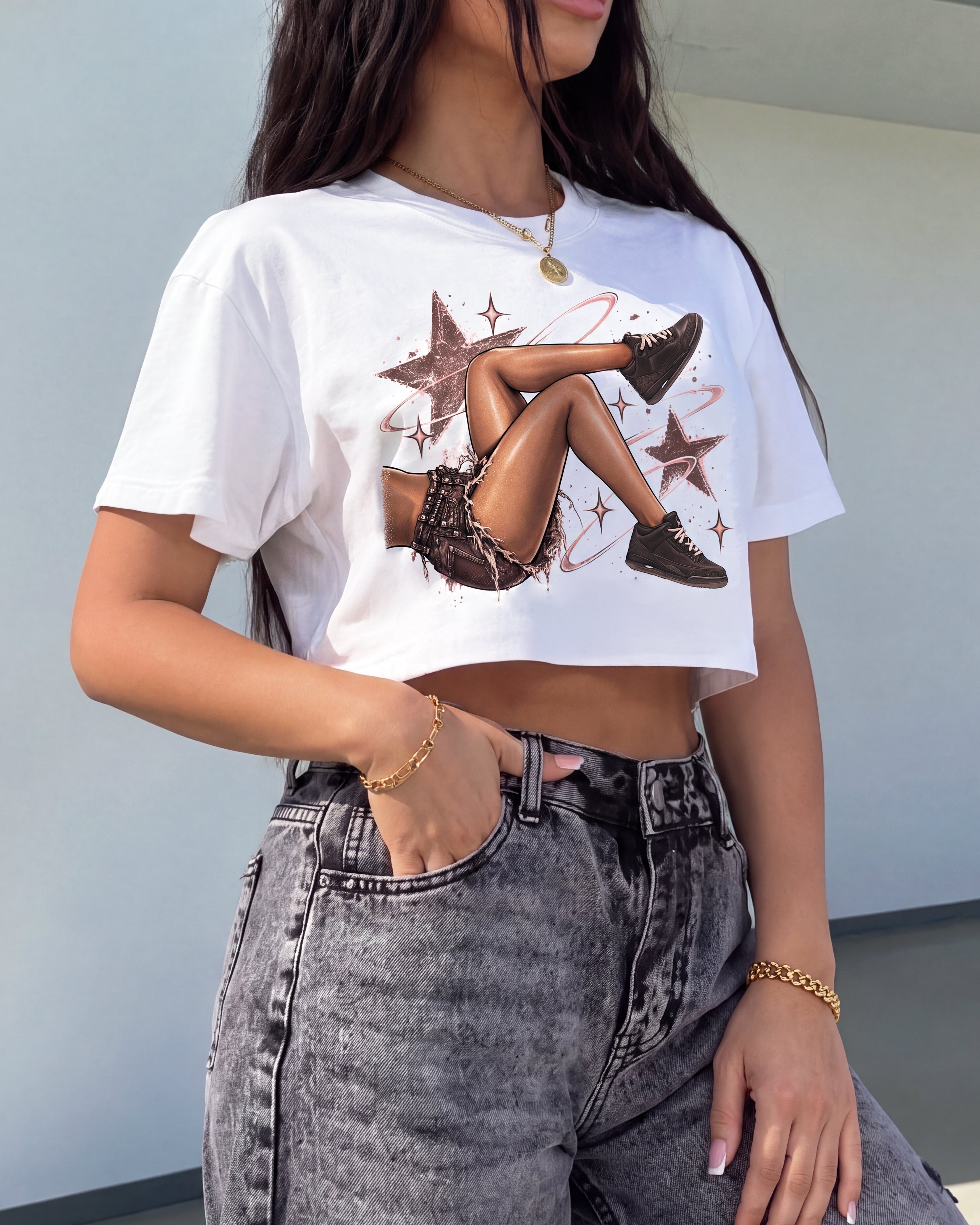 A woman wearing a white cropped graphic t-shirt with a vintage-style illustration of a woman's legs and brown stars, paired with high-waisted gray acid-wash jeans.