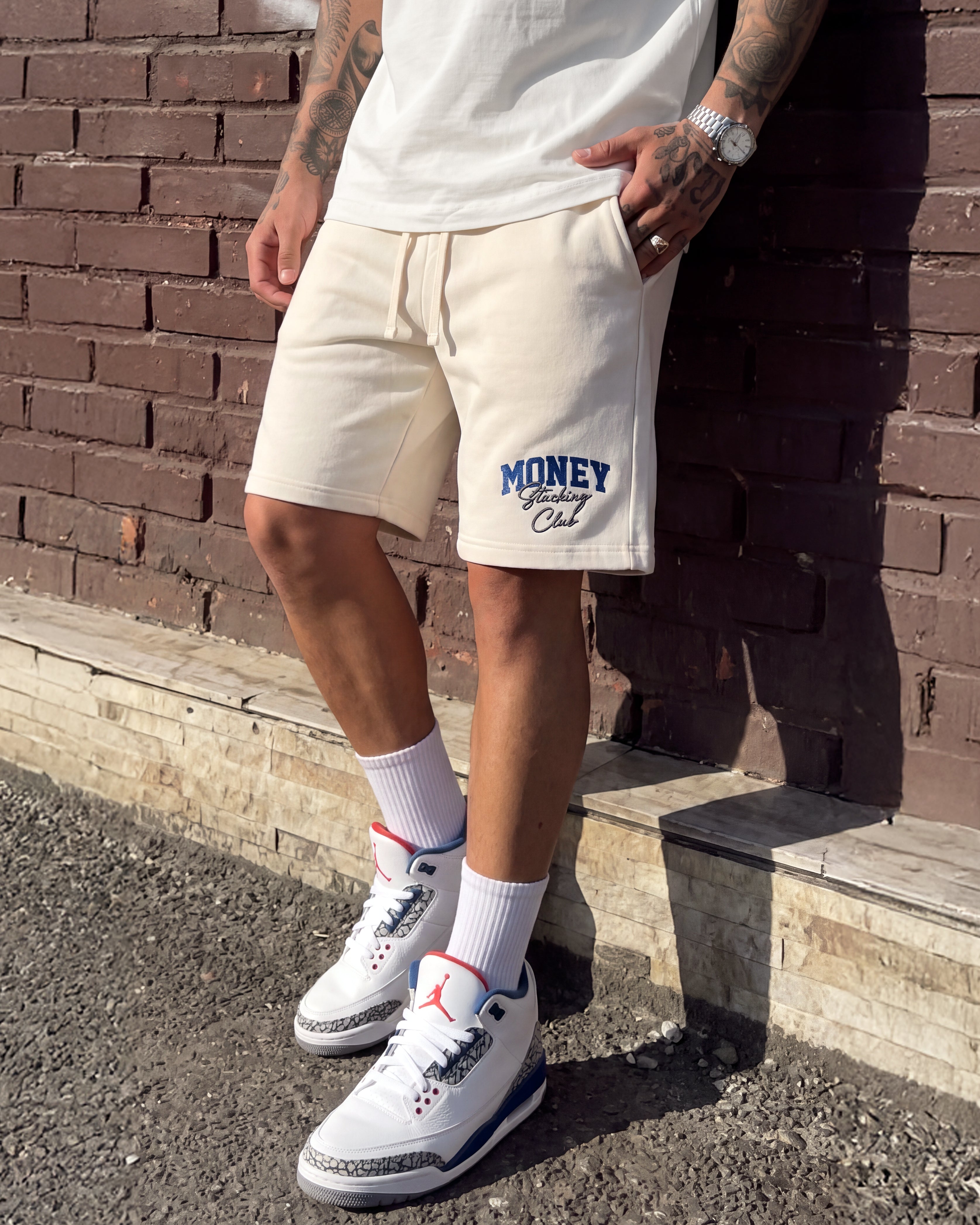 A person wearing off-white athletic shorts with a blue 'MONEY God Call' graphic on the left leg, paired with white sneakers and white crew socks, leaning against a brown brick wall.