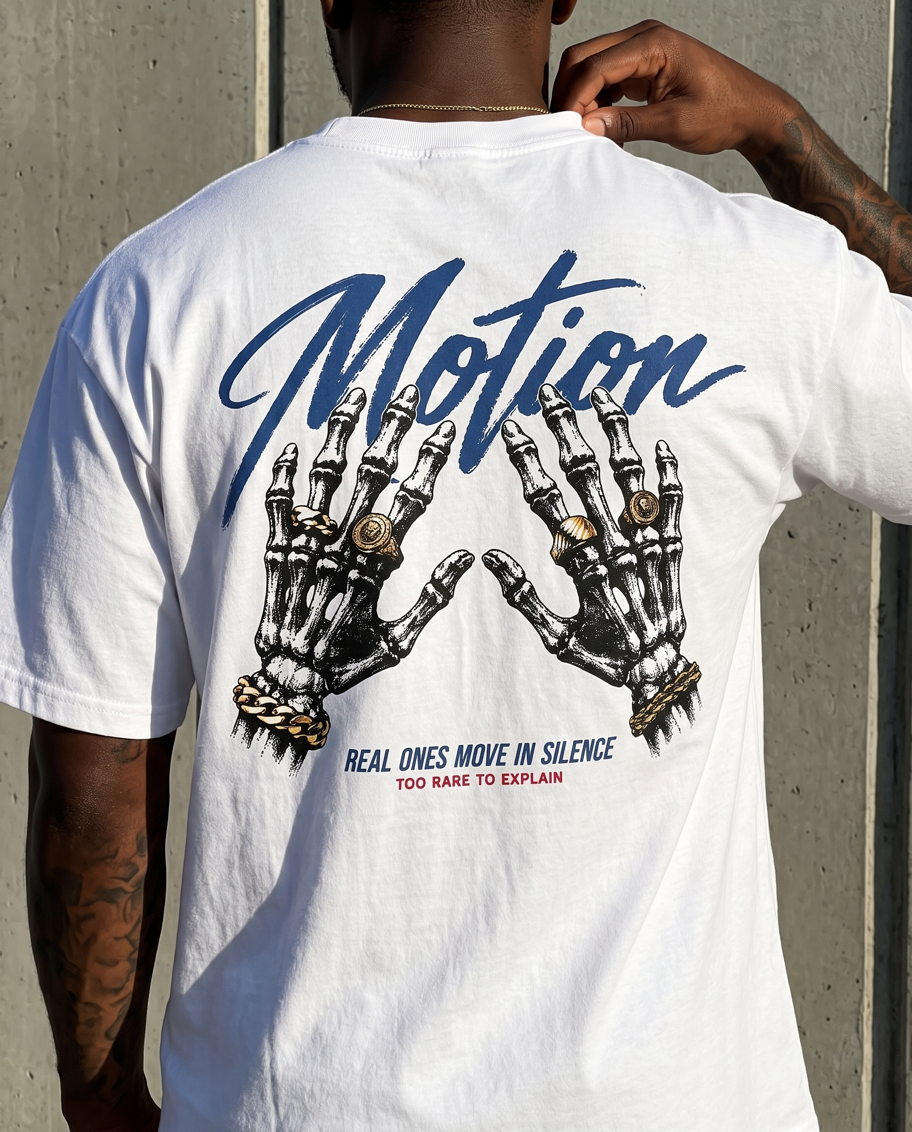 A man viewed from behind wearing a white t-shirt with a large graphic print on the back. The graphic features two skeletal hands with gold rings and bracelets under the word 'Motion' in blue script, with the text 'REAL ONES MOVE IN SILENCE TOO RARE TO EXPLAIN' below.