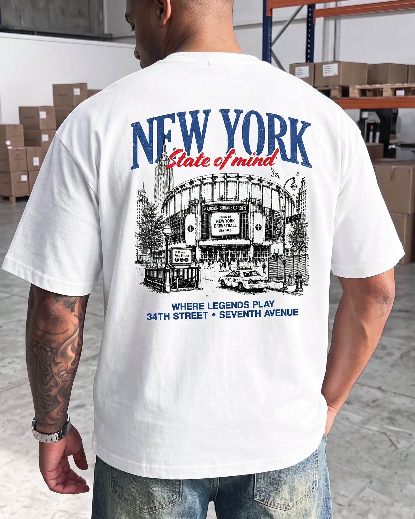 A man viewed from behind wearing a white graphic t-shirt with a detailed black and blue print of Yankee Stadium in New York City. The text reads 'NEW YORK State of mind' and 'WHERE LEGENDS PLAY 34TH STREET • SEVENTH AVENUE'.