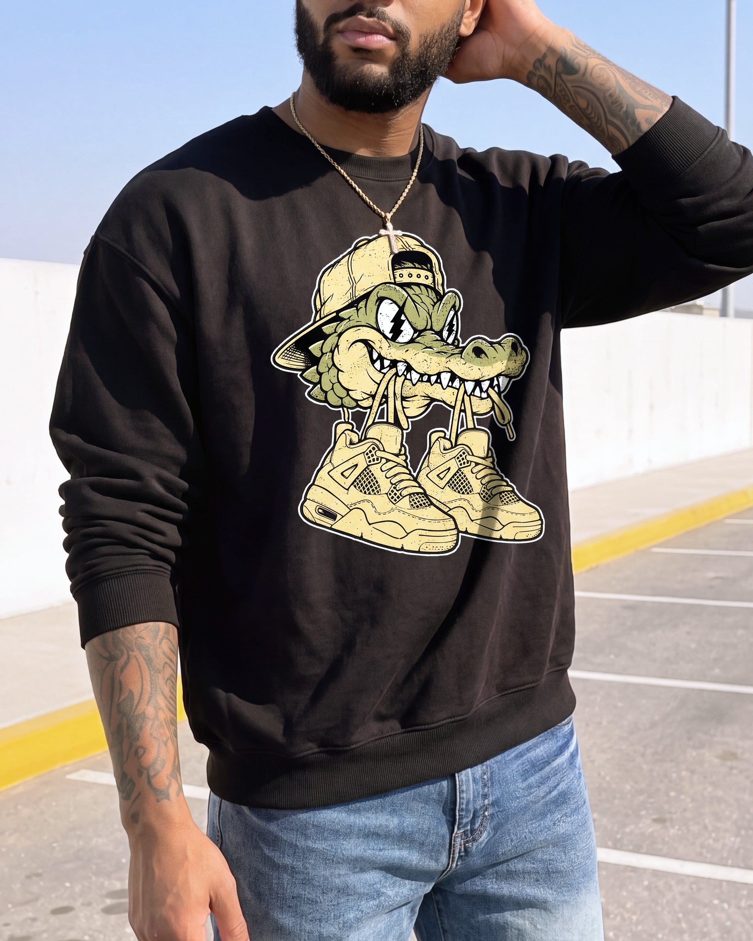 A man wearing a black crewneck sweatshirt with a cartoon alligator graphic wearing a tan cap and beige sneakers, paired with blue jeans, photographed outdoors in a parking lot.